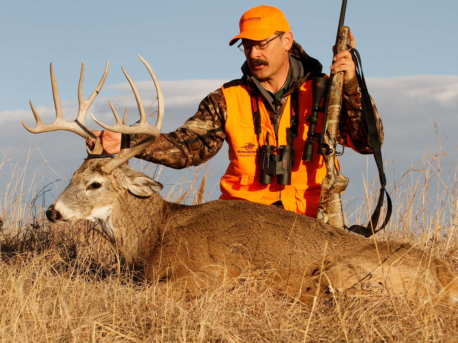 A hunter in camo and orange kneels behind a whitetail buck and holds its antlers.