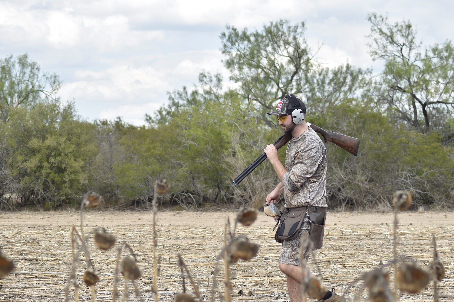 A hunter walks through a large open field with a shotgun over his shoulder.