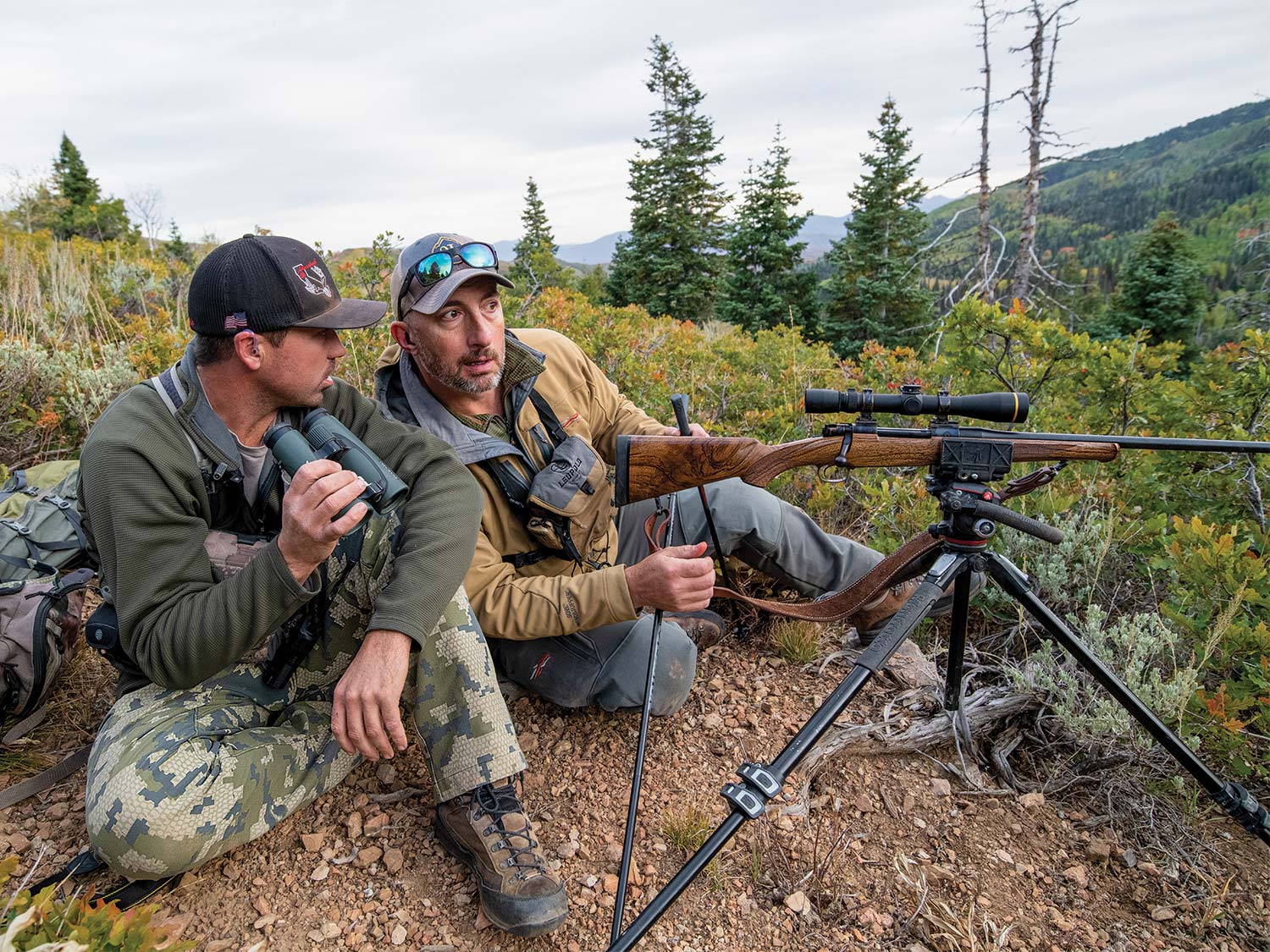 Two hunters sit on a hillside with a rifle on a tripod and scout through binoculars.