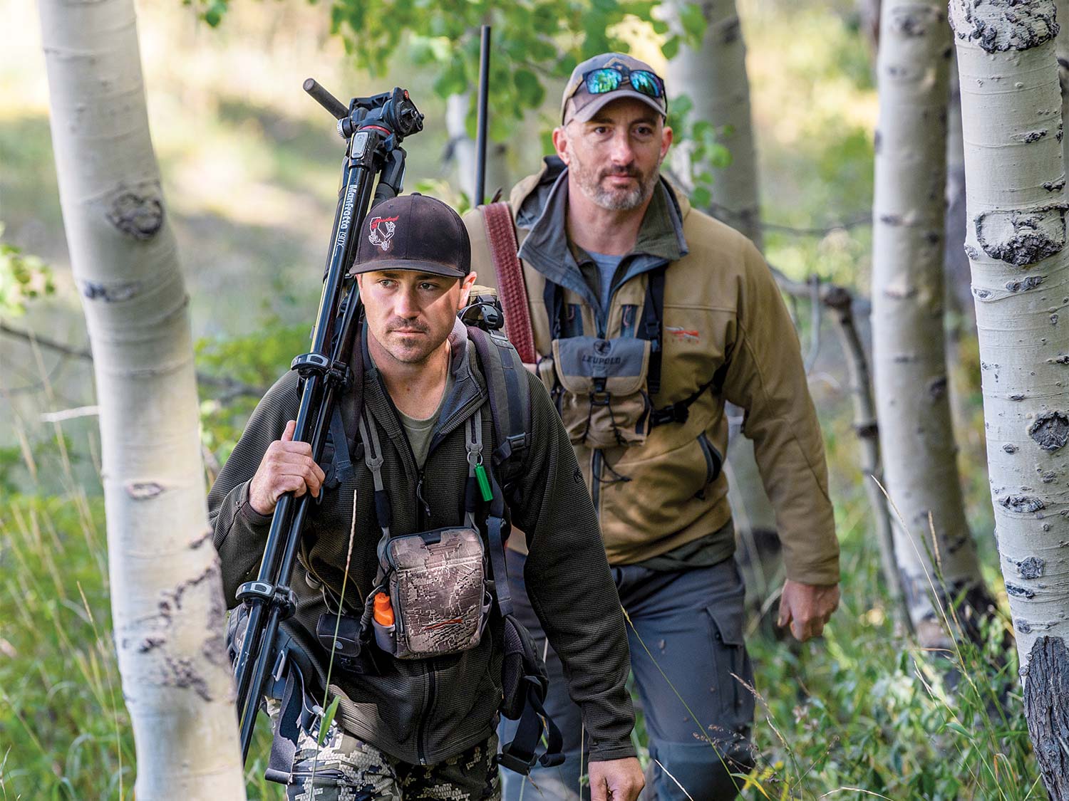 Two hunters hike through the woods and carry their gear with them.