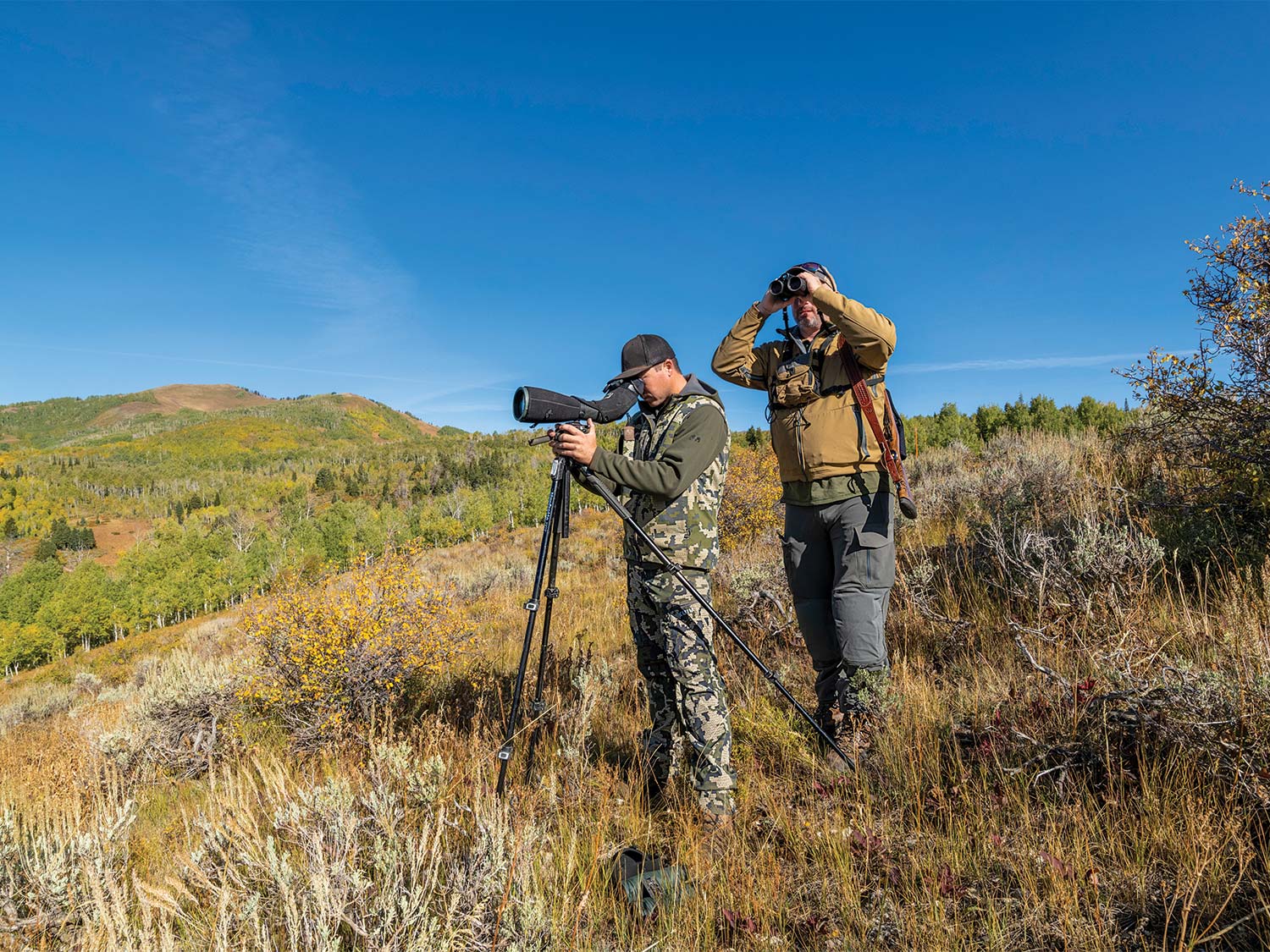 Two hunters stand on a hillside and scout the terrain using spotting scopes and binoculars.