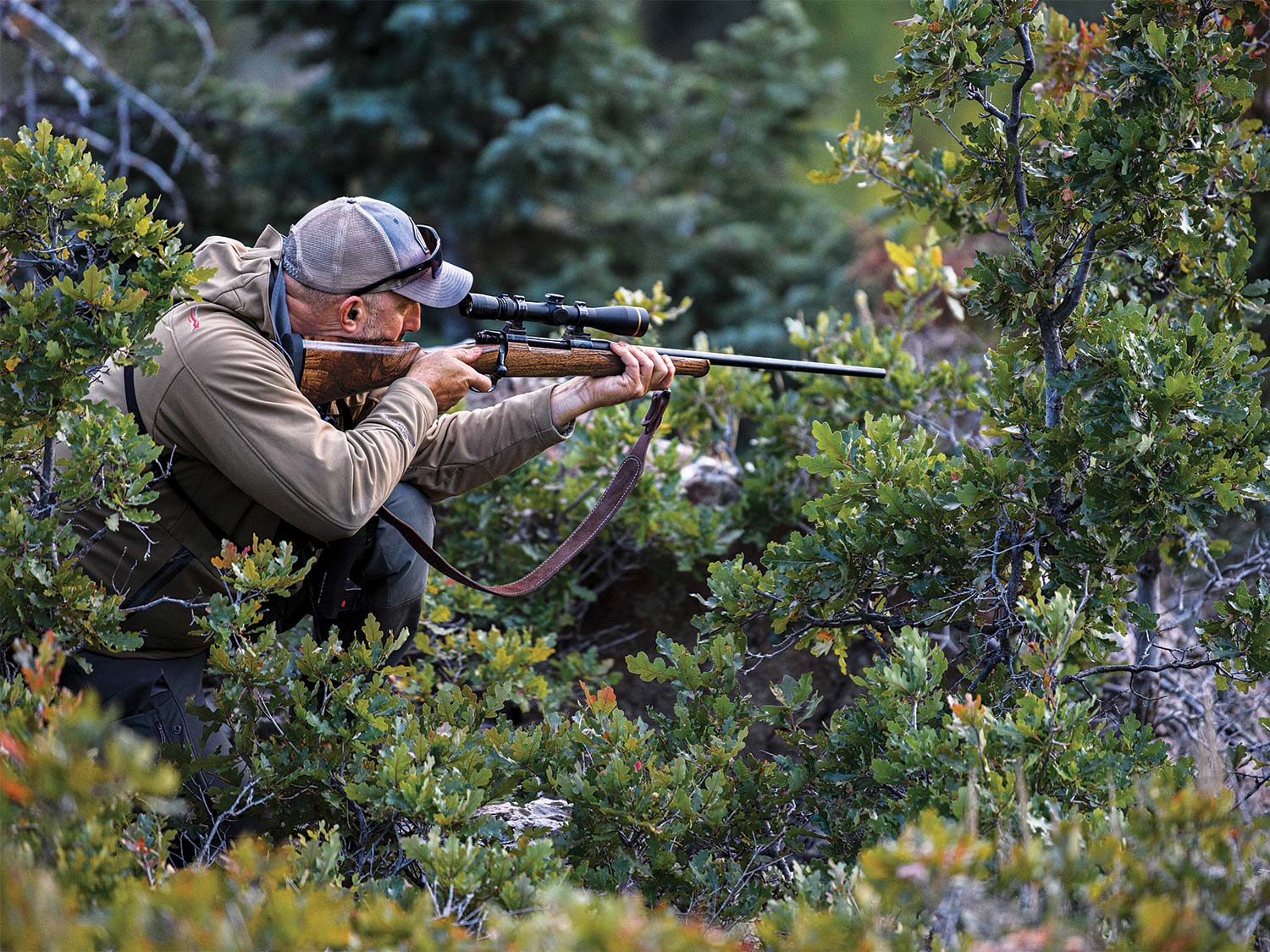 A hunter stands amongst brush and cover while looking through a riflescope and holding a rifle against his shoulder, ready to fire.