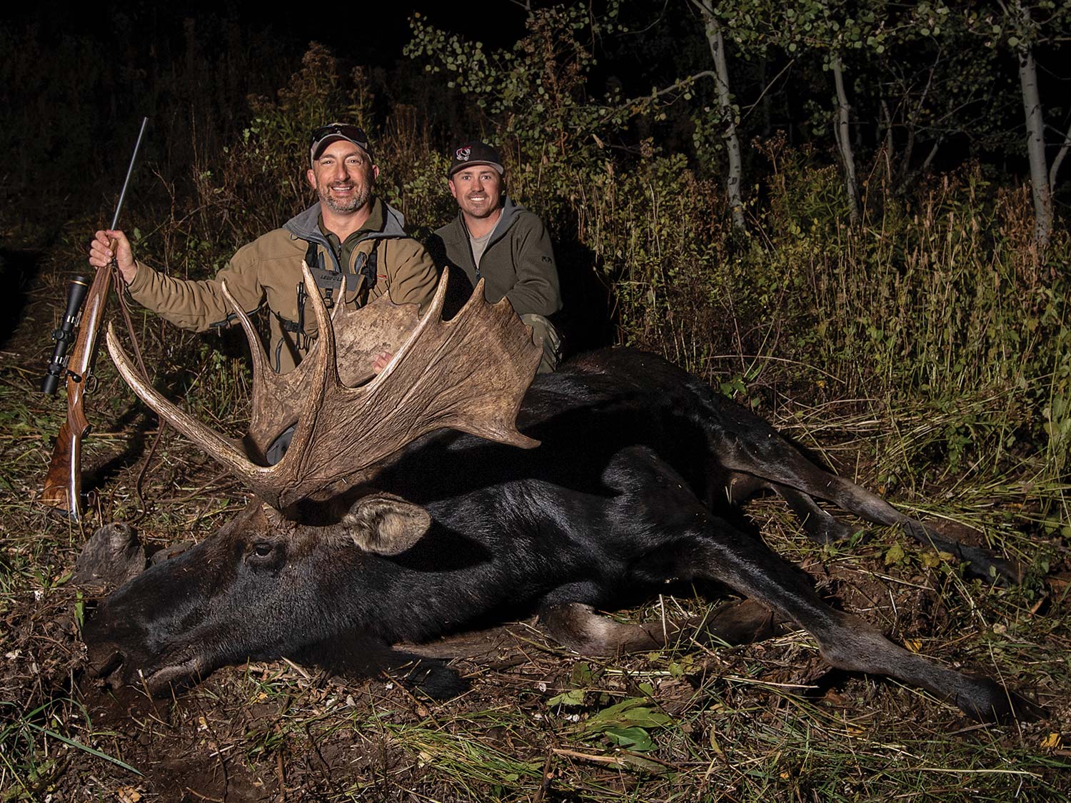 Two hunters kneel behind a large bull moose at night.