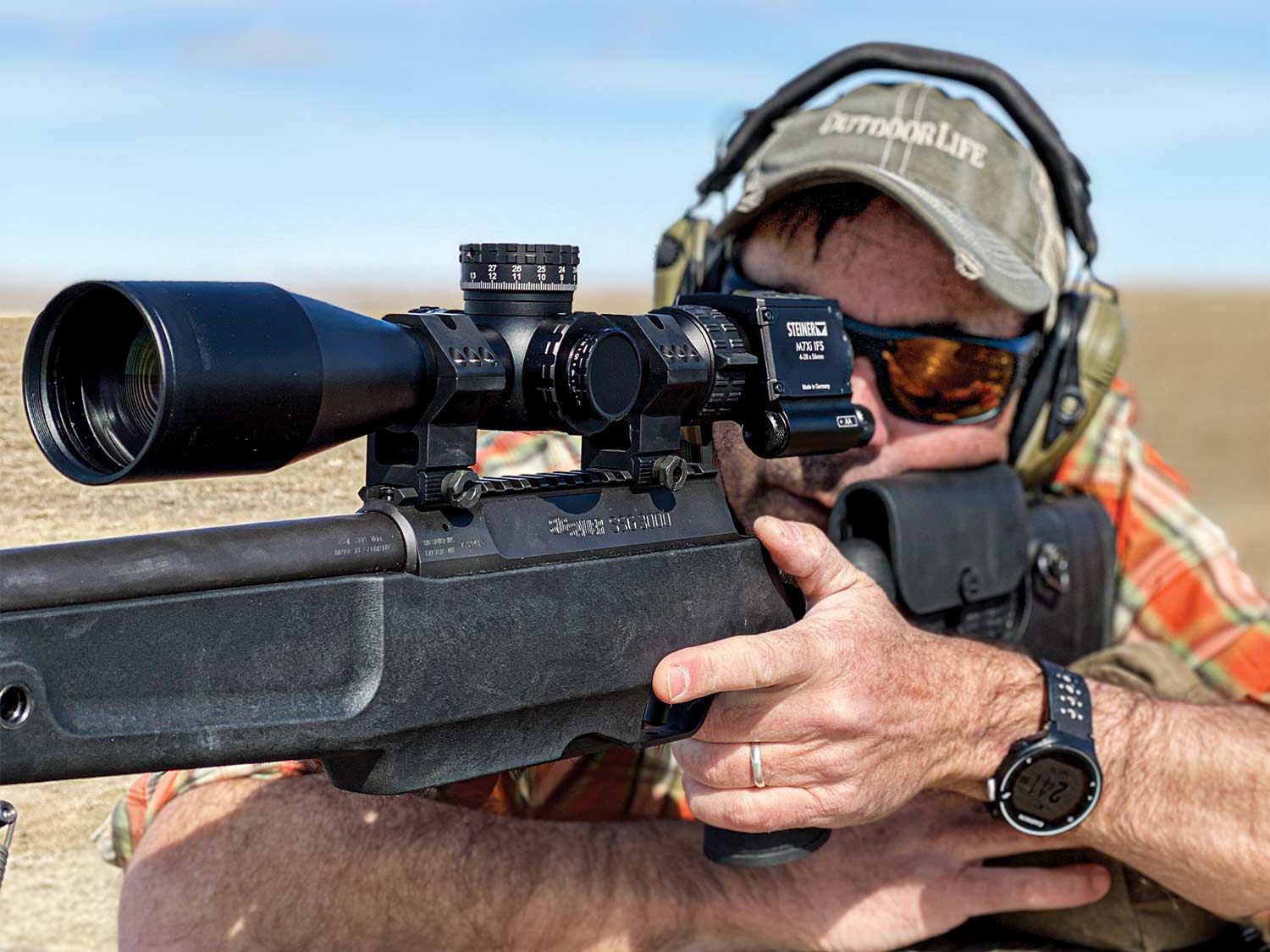 A man in a ball cap aims a rifle and looks through the rifle scope.