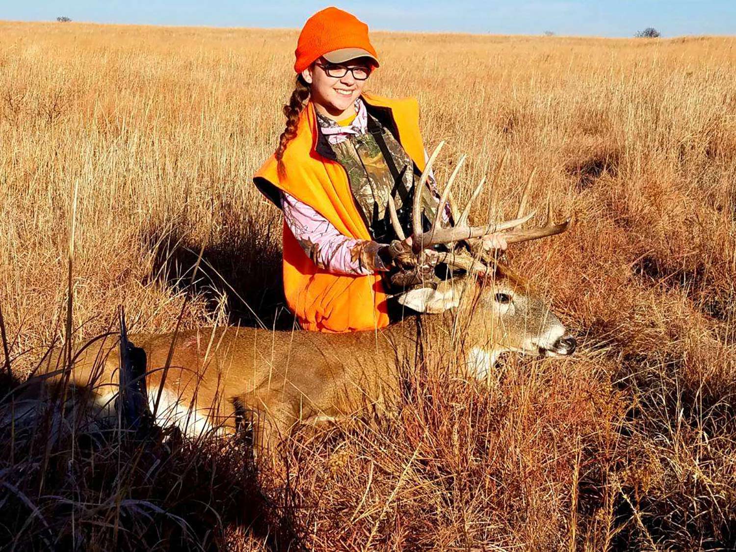 A young girl clad in hunter orange kneels behind a whitetail buck in a large open field and holds its head up by the antlers.