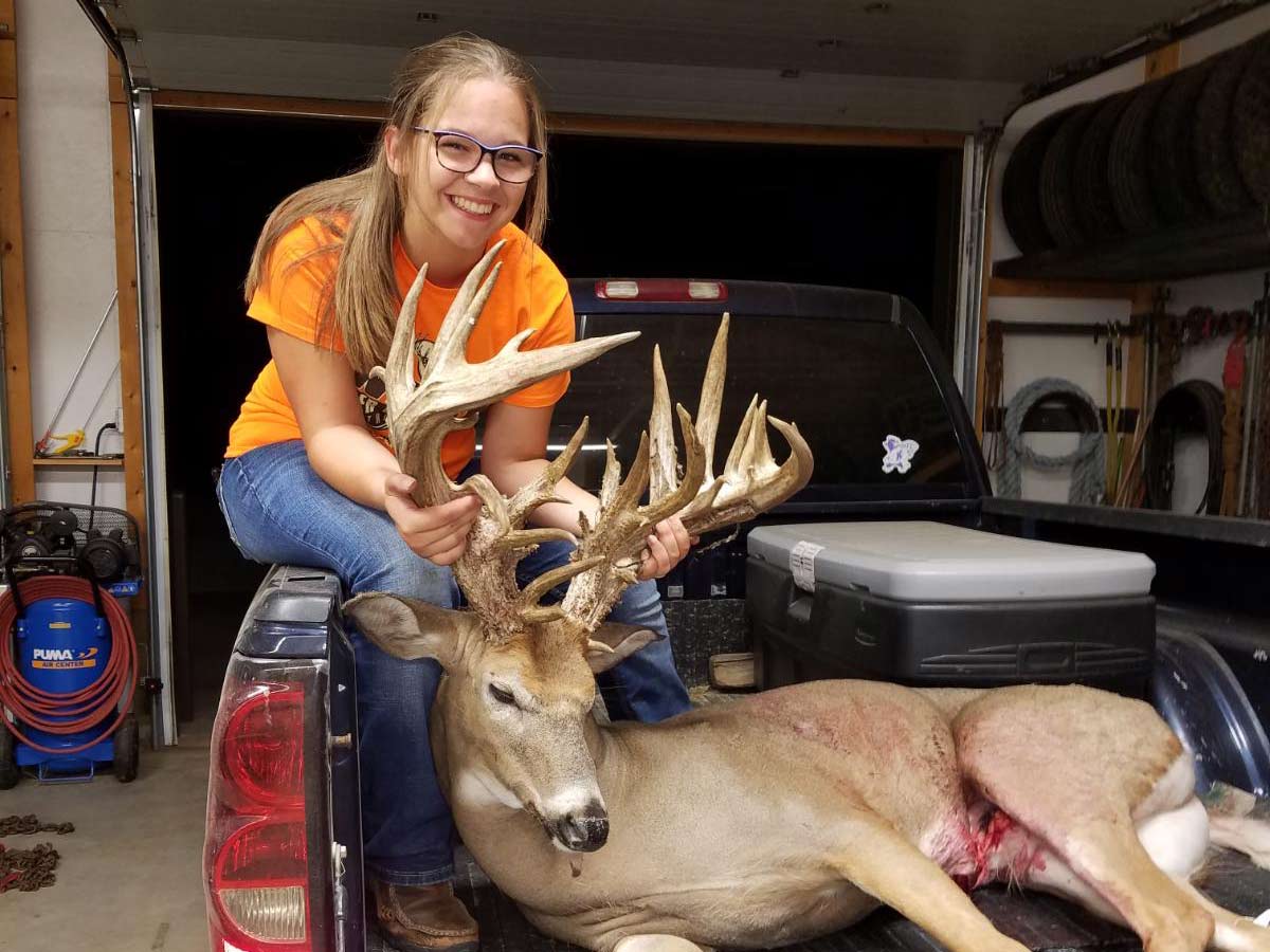 A young girl sits on a truck bed behind a dead whitetail deer and holds up its head by the antlers.