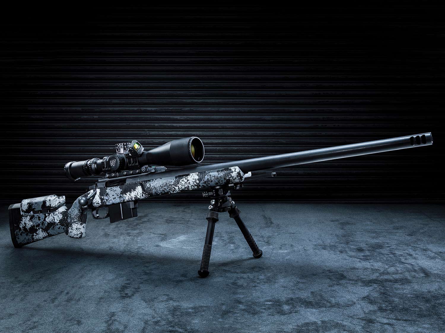 A black and grey camo-patterned rifle equipped with a riflescope and resting on shooting bipods.