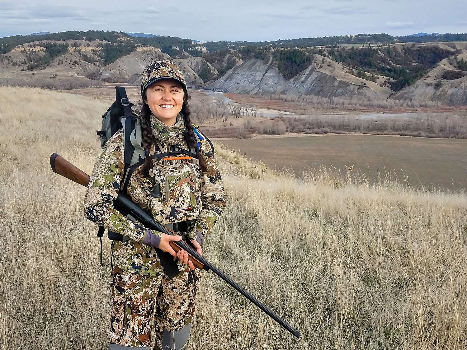 A woman wearing full camo and holding a shotgun stands in a large midwestern plain with hills and mountains in the background.