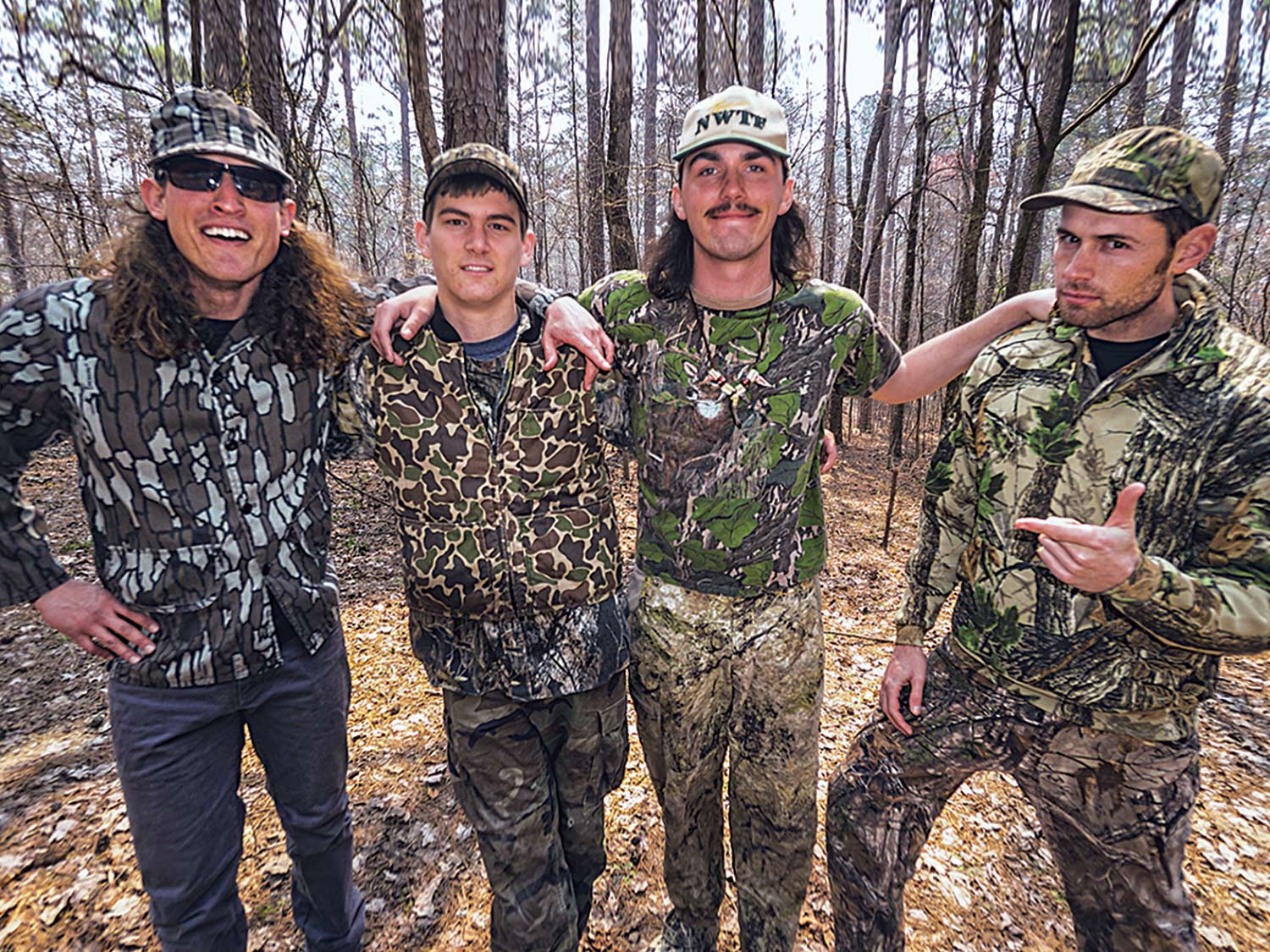 Four hunters in full  camo stand out in the woods, posing and smiling together.