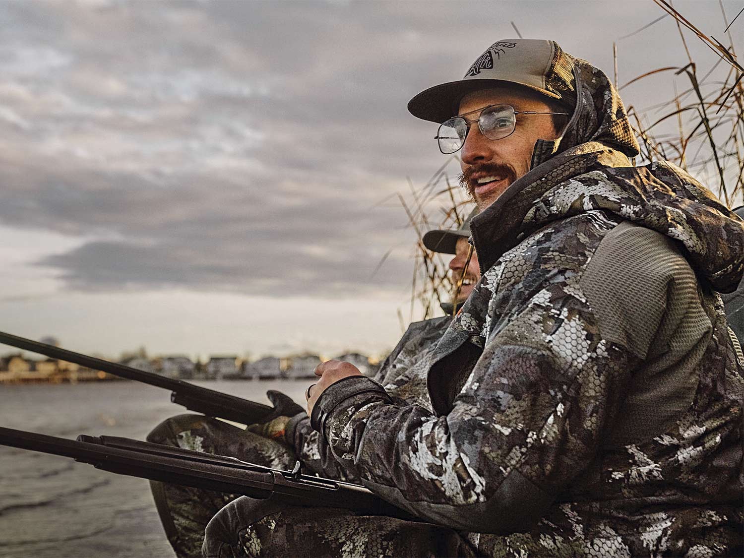 A man in glasses and wearing full camo sits by the waterside with a shotgun in his lap.