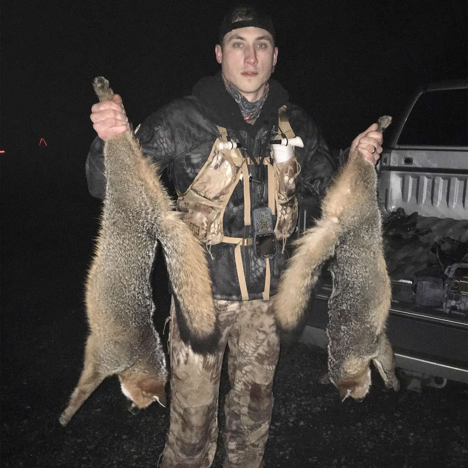 A man in hunting gear holds up two grey foxes at night.