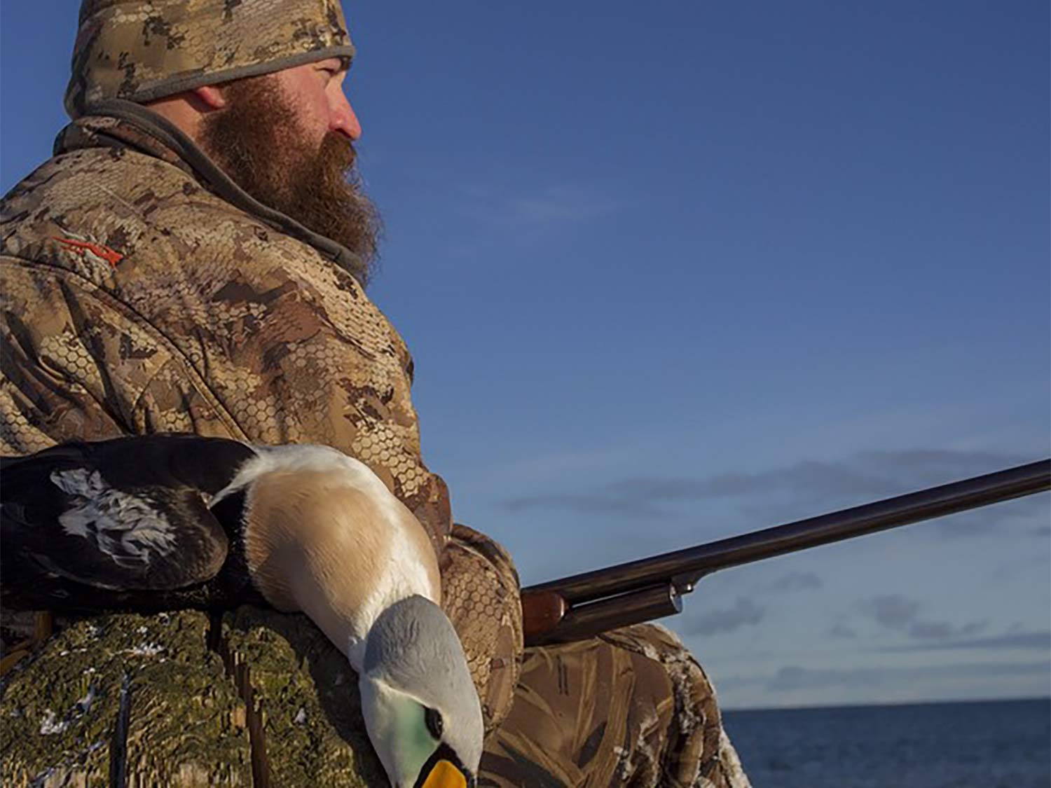 A hunter sits with a shotgun on his lap next to a duck.