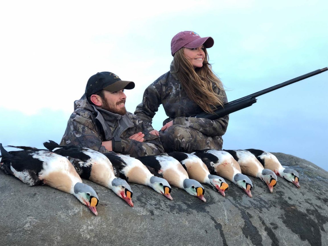 Two hunters, man and woman, sit on a rock next to a limit of king ducks.
