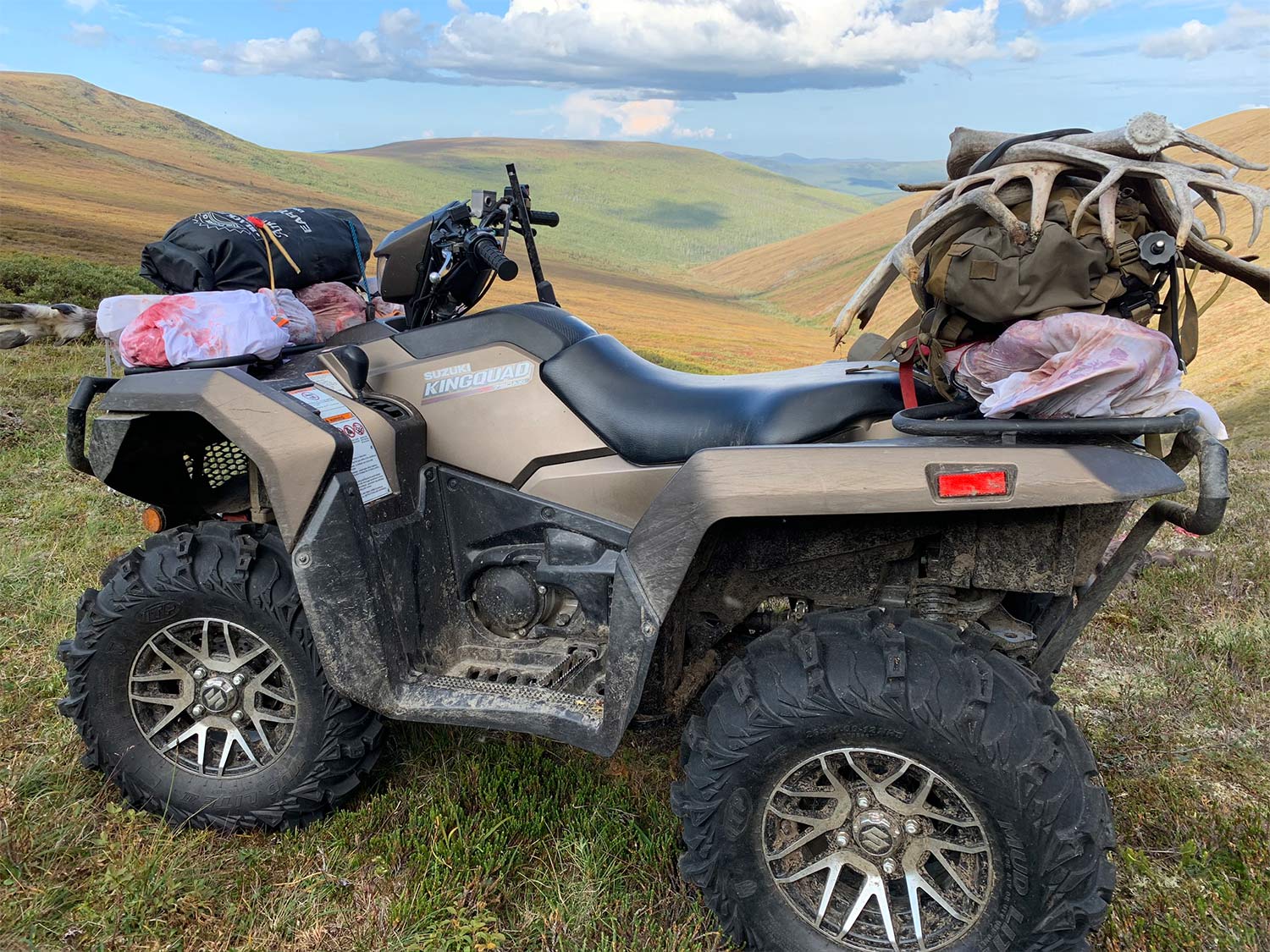 A four-wheeled ATV loaded with hunting gear sits atop a hill with a sprawling view of hills and plains in the background.
