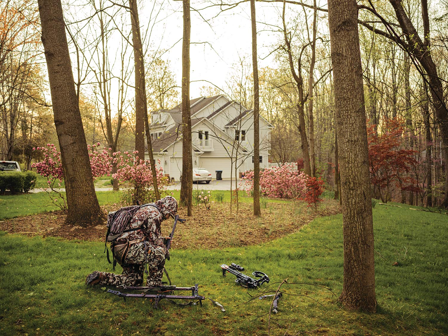 A hunter in full camo prepares hunting gear and tree stands in a suburban back yard.