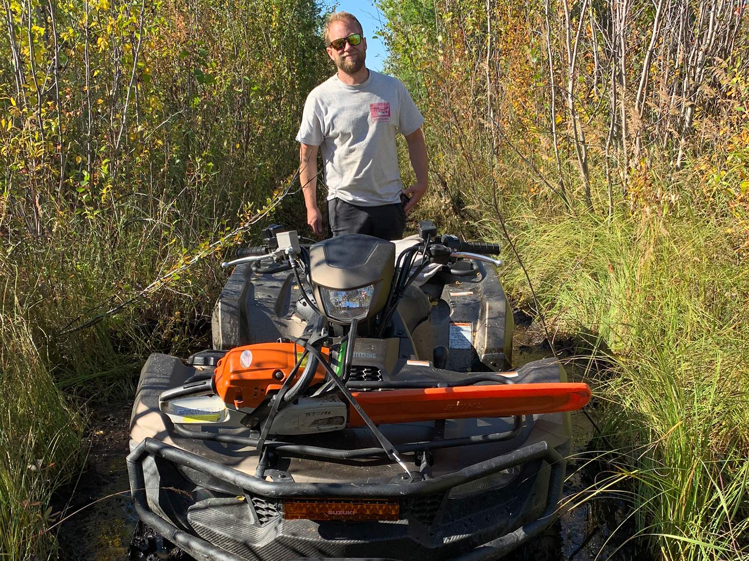 A man stands behind a four-wheeled ATV in very dense brush and woods, on a hunting trail.