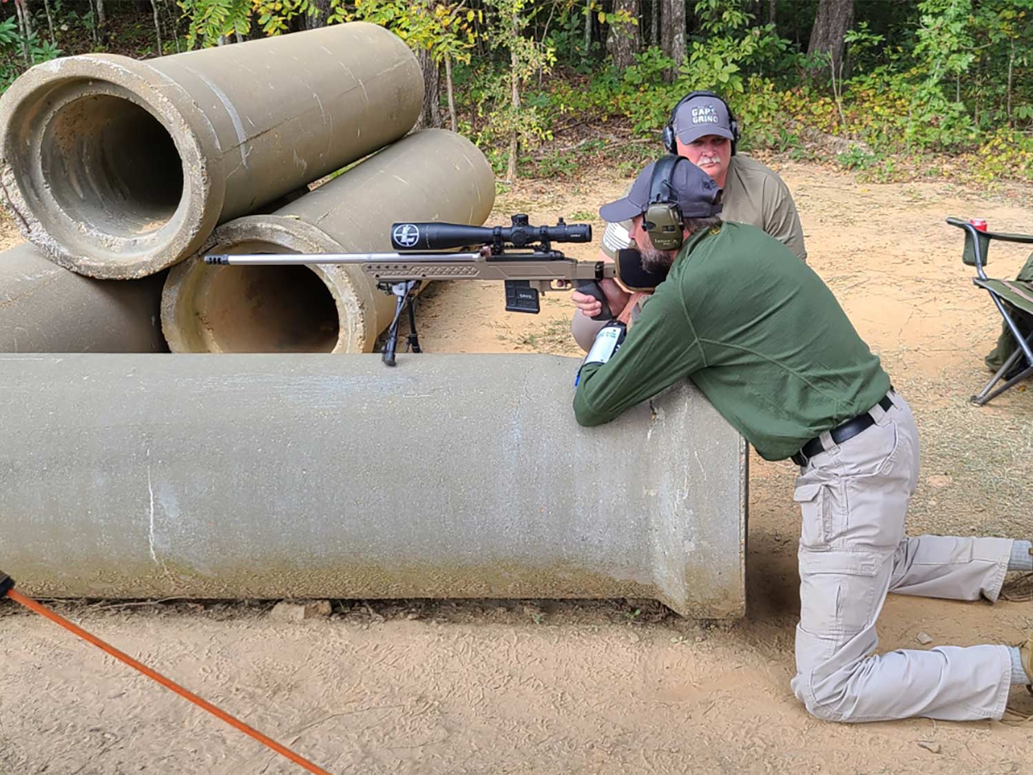 A man leans over a pipe and aims a rifle while glancing through a riflescope.