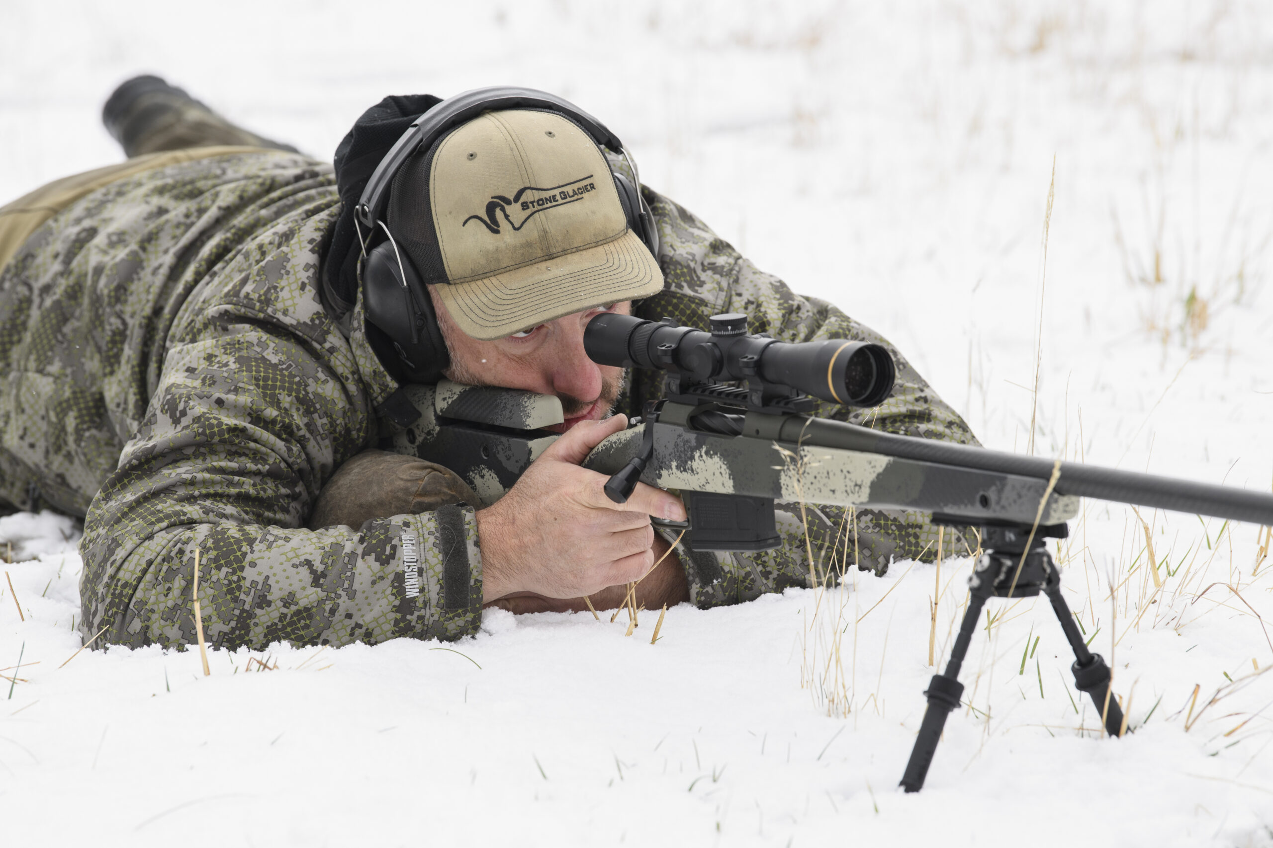 A hunter in camo and a green hat lies prone on the ground behind a Springfield Waypoint rifle, in conjunction with a bipod.