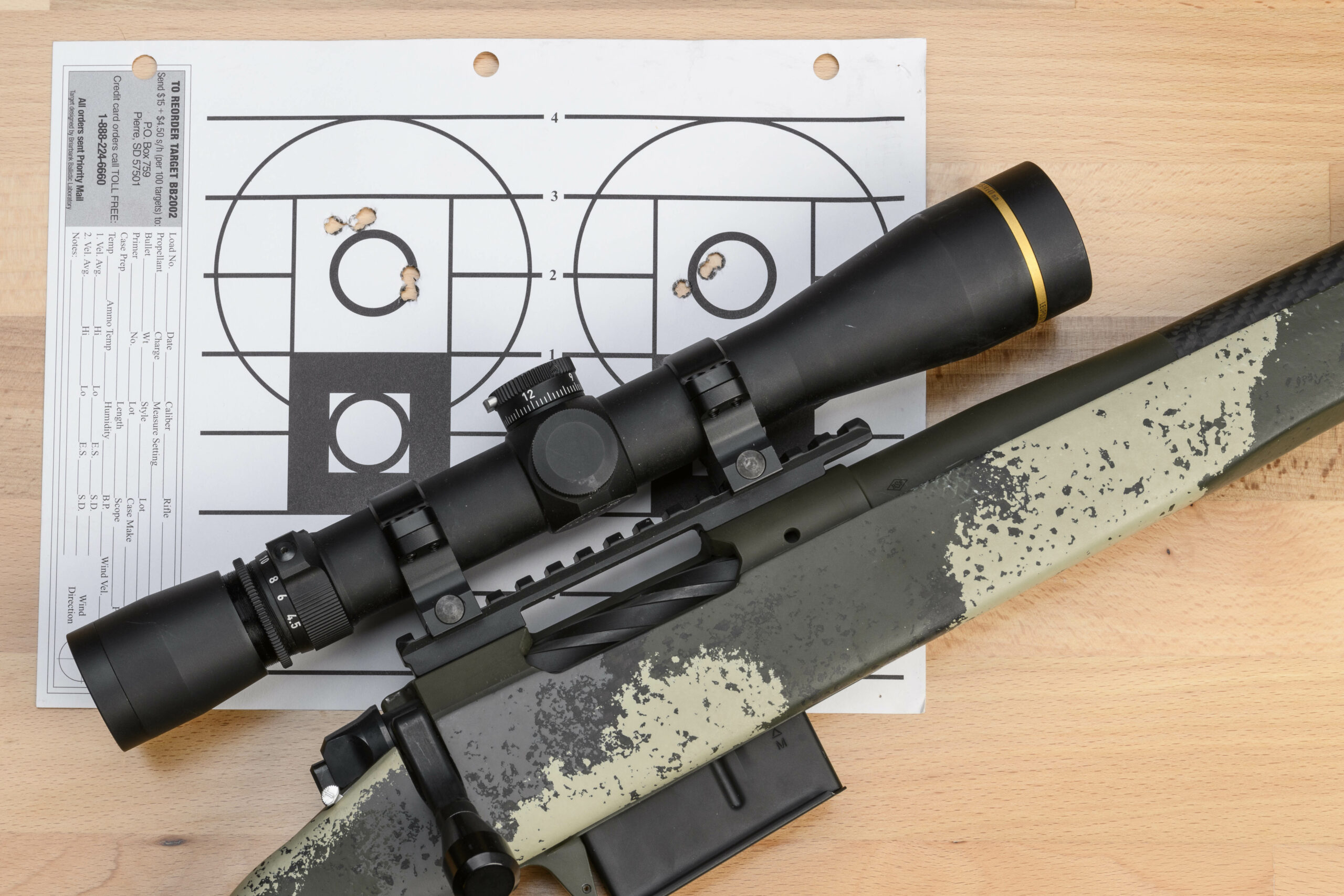 A camo-stocked bolt-action rifle, topped with a black scope, lying on top of a paper target with two zeroing groups.