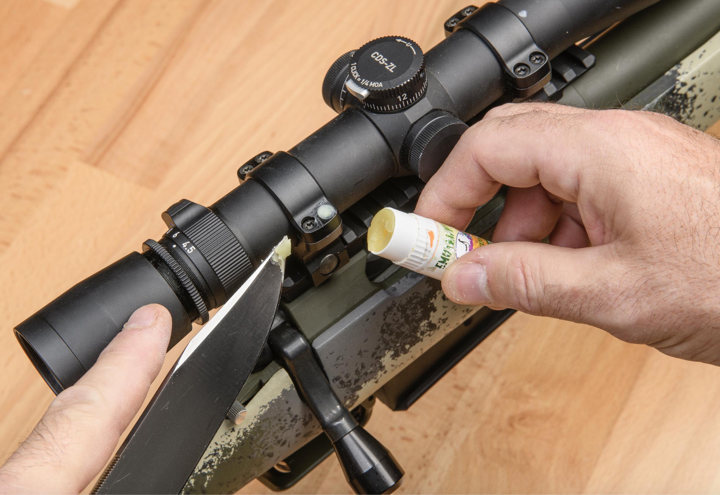 A pair of hands holds a chapstick tube and a knife-tip coated in chapstick while applying it to one of the screw heads on the riflescope bases.