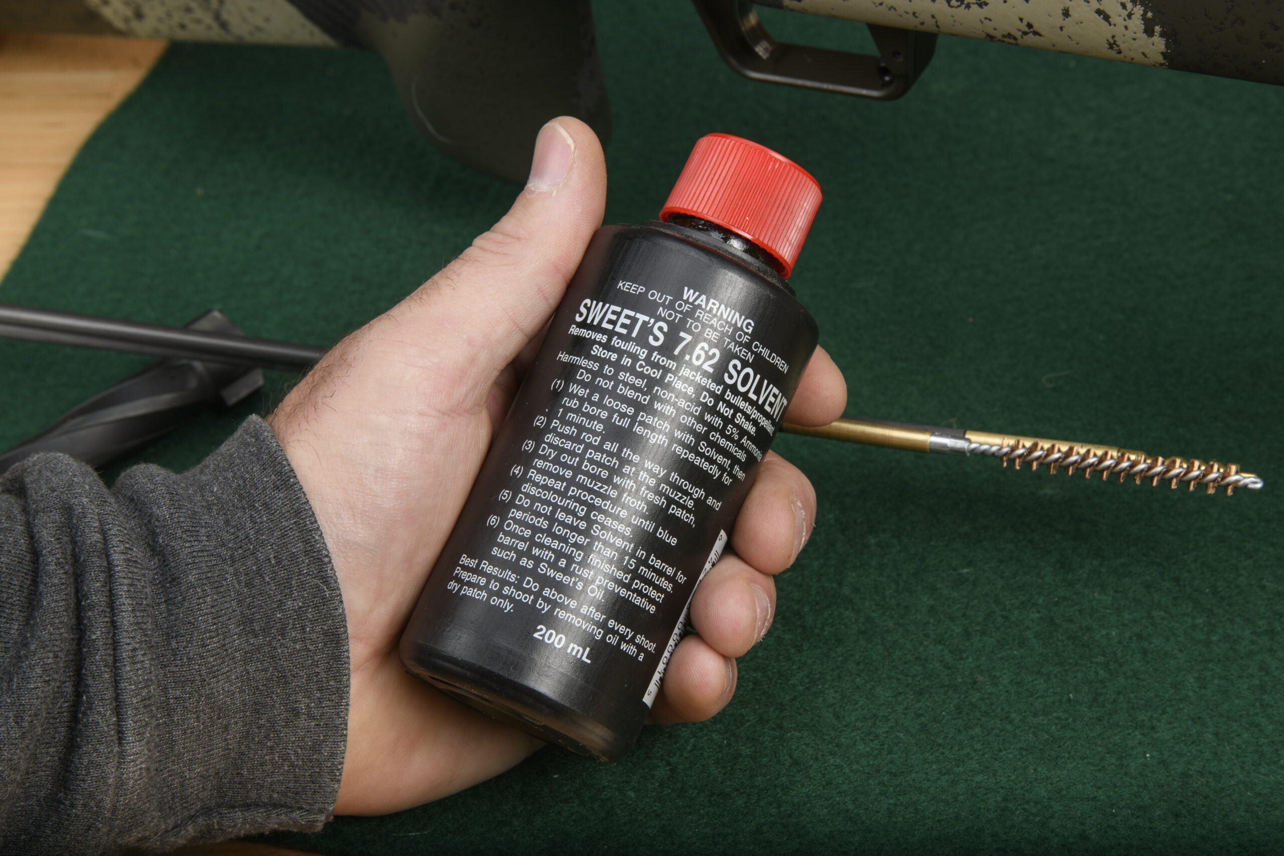 A hand holding a black bottle with a red cap and the words "Sweet's 7.62 Solvent" on top of a green felt gun cleaning pad.