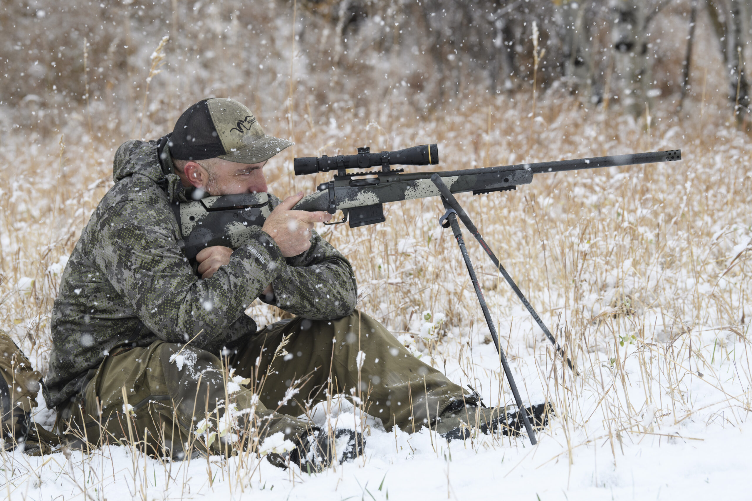 Hunter sits behind a Springfield Waypoint rifle with a pair of shooting sticks in the snowy brush.