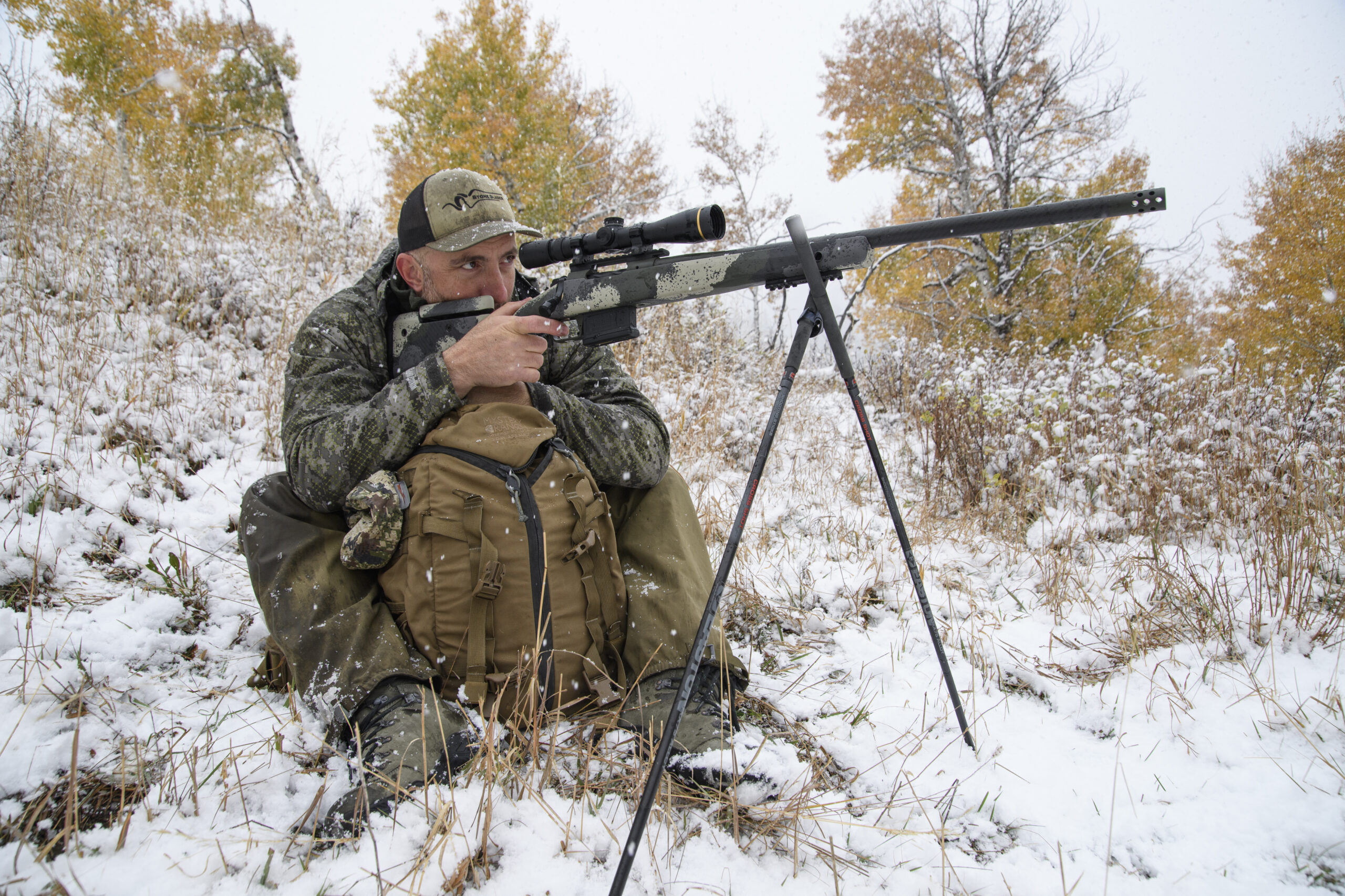 A hunter shoots his rifle off sticks on a snowy hillside while using a pack to steady himself.