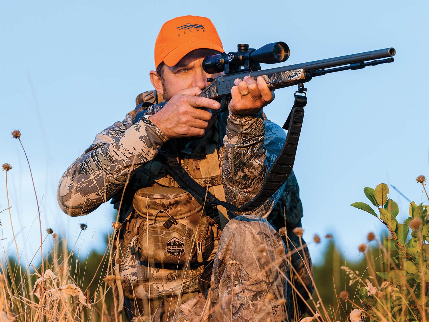 A hunter in full camo and orange cap holds a rifle to their shoulder while aiming, preparing to fire.