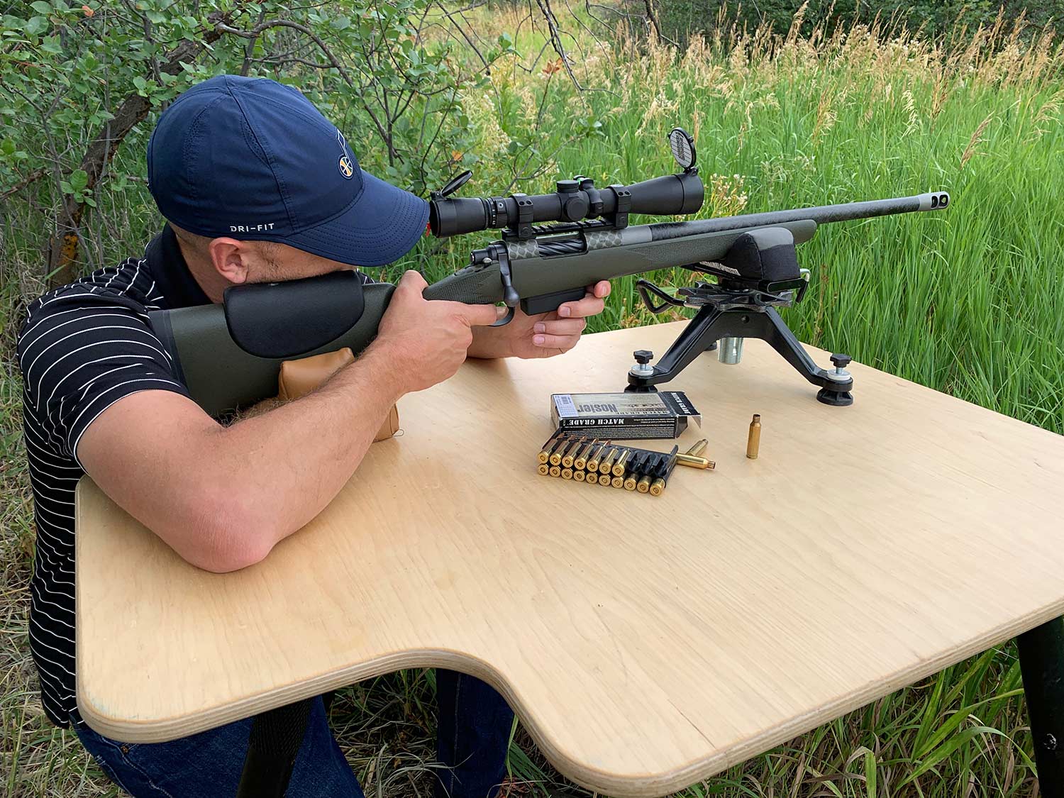A man aiming a rifle propped on a table and shooting bipod.