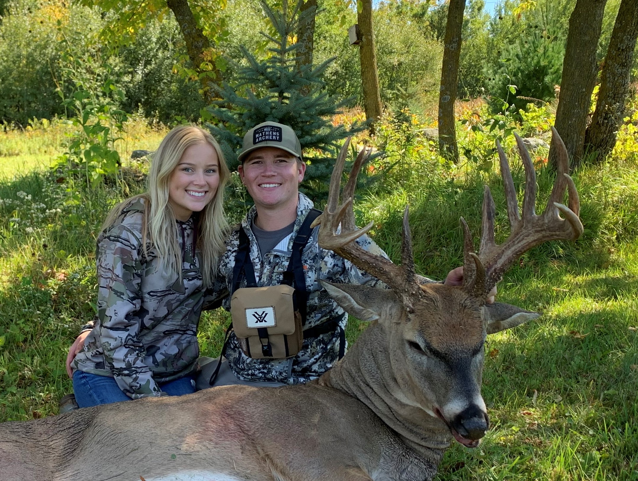 A smiling young man and woman in camo shirts sit behind a big whitetail buck.