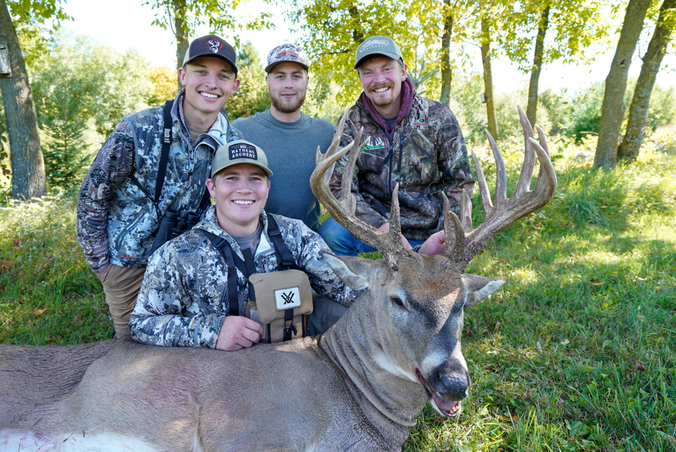 Four young smiling hunters sit and stand behind a big whitetail buck.