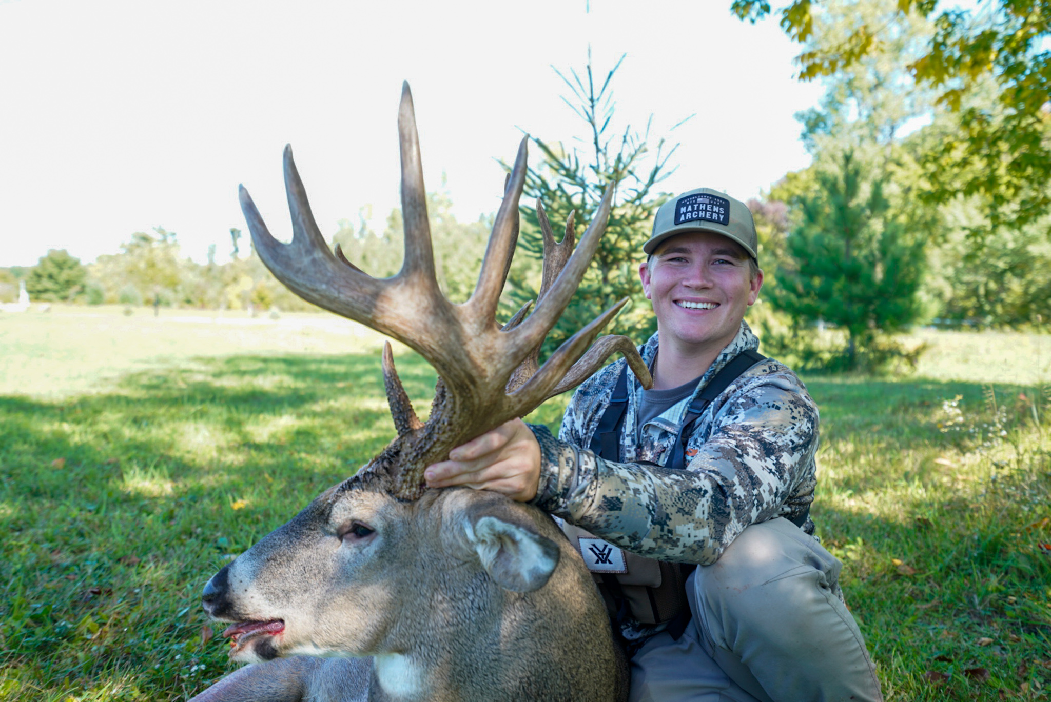 A grinning young hunter in a camo shirt holds the head of a whitetail buck, whose antlers are in profile.