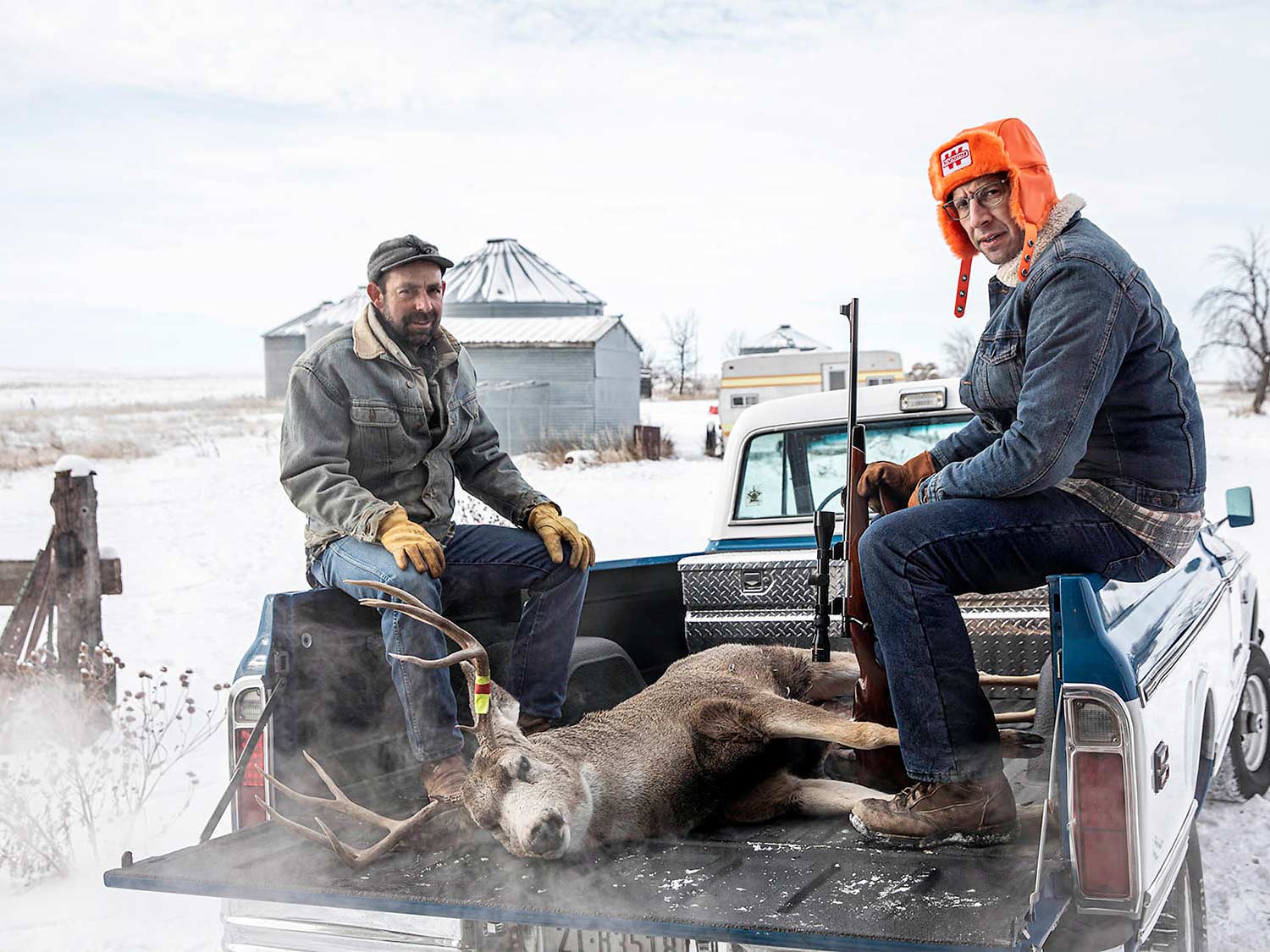 Two hunters sit and pose in the bed of a truck next to a mule deer.