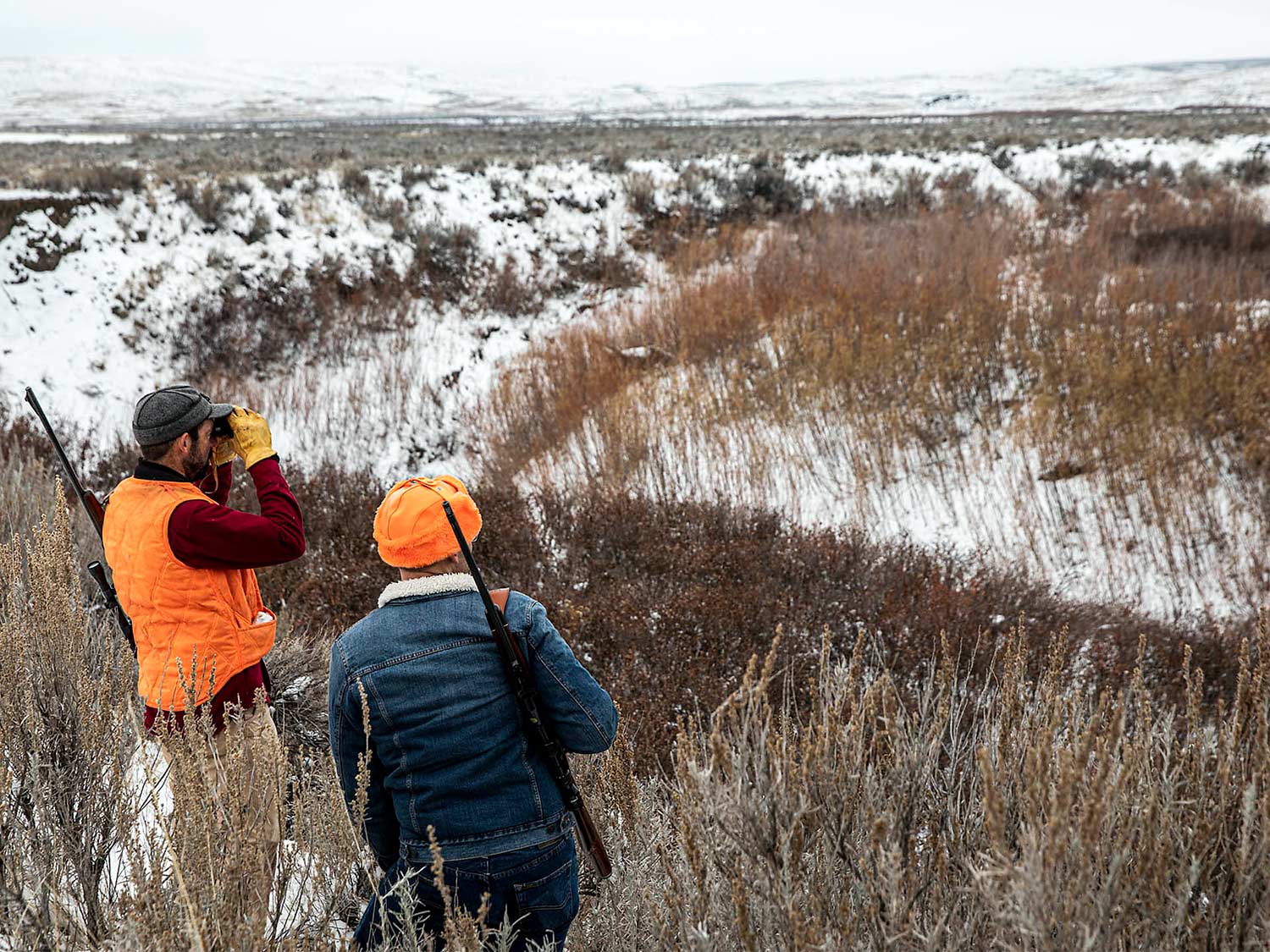 Two hunters scout an open, snow covered plain.
