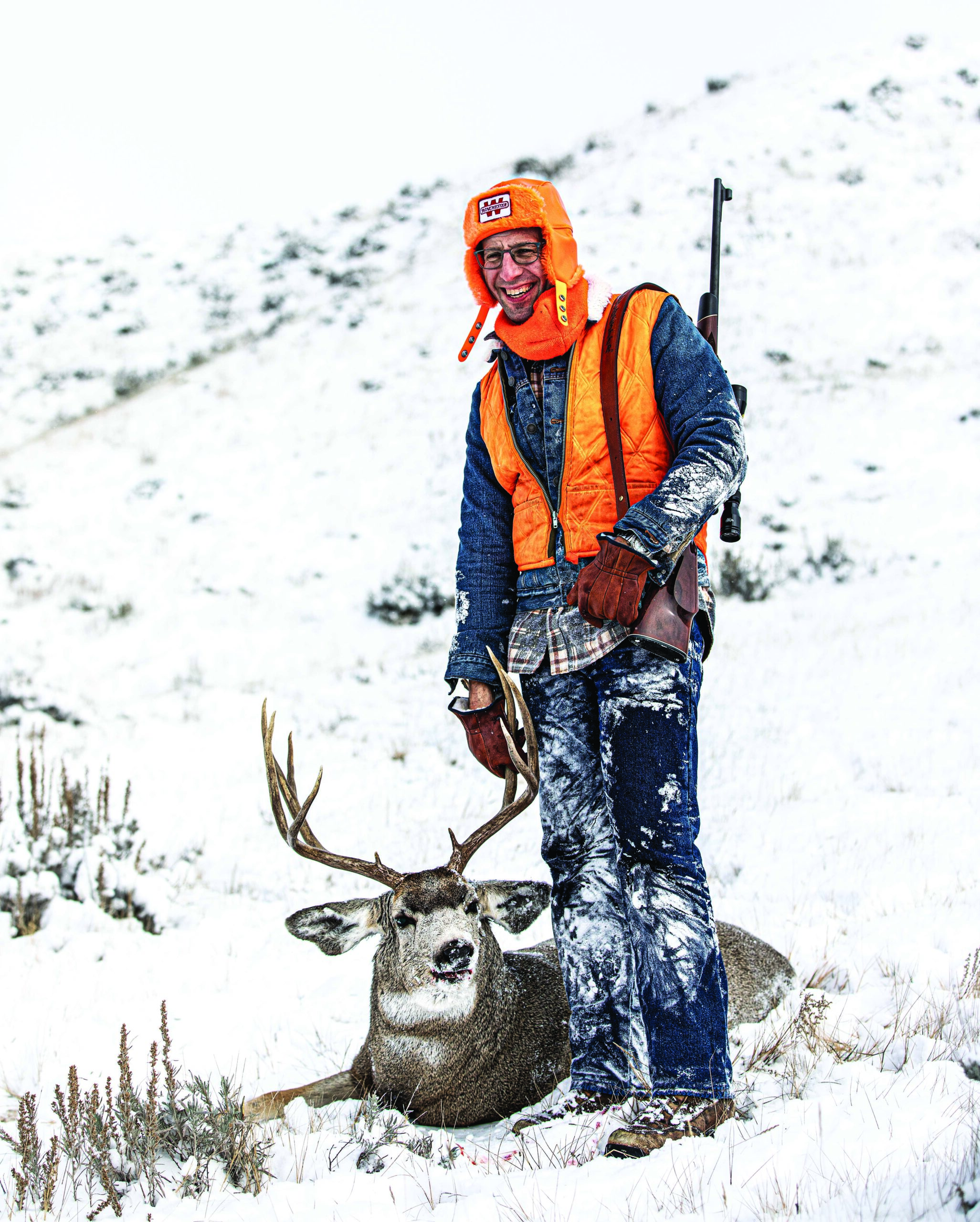 A hunter in denim and blaze orange with a snow Montana mule deer buck.