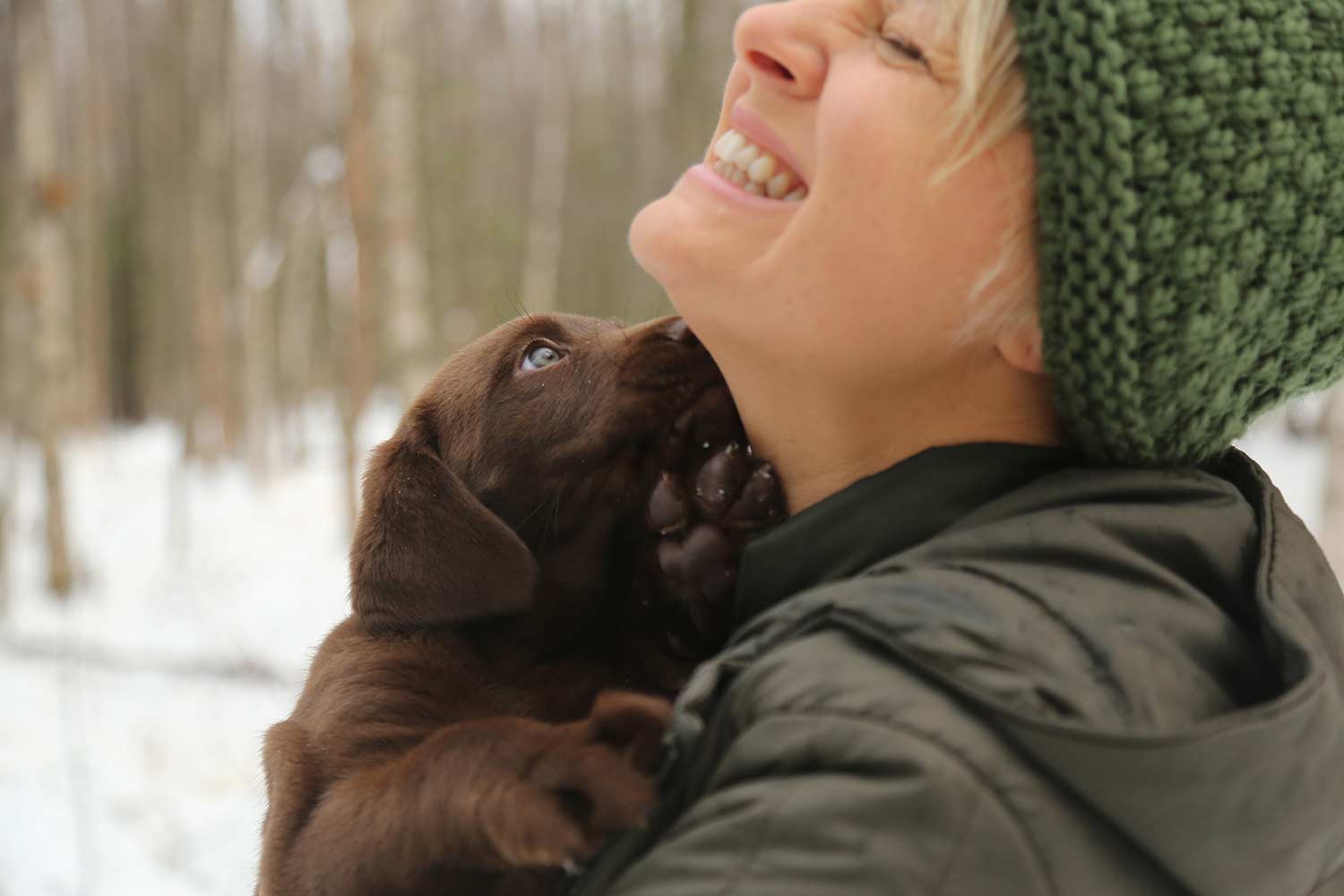 A woman hunter holds a chocolate lab puppy while standing in the snow.