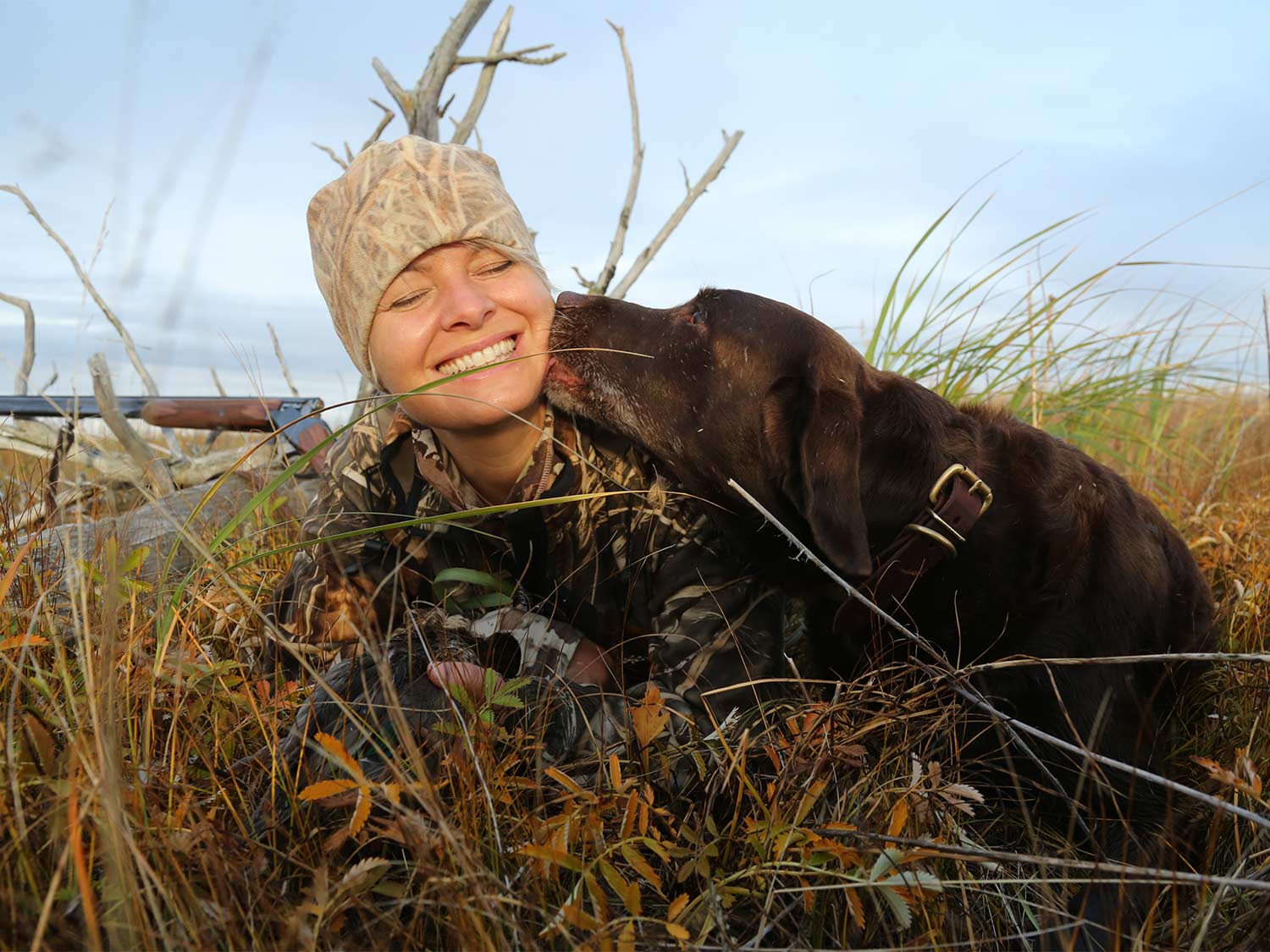 A woman in full hunters camo sits in a field while a chocolate lab licks her face.