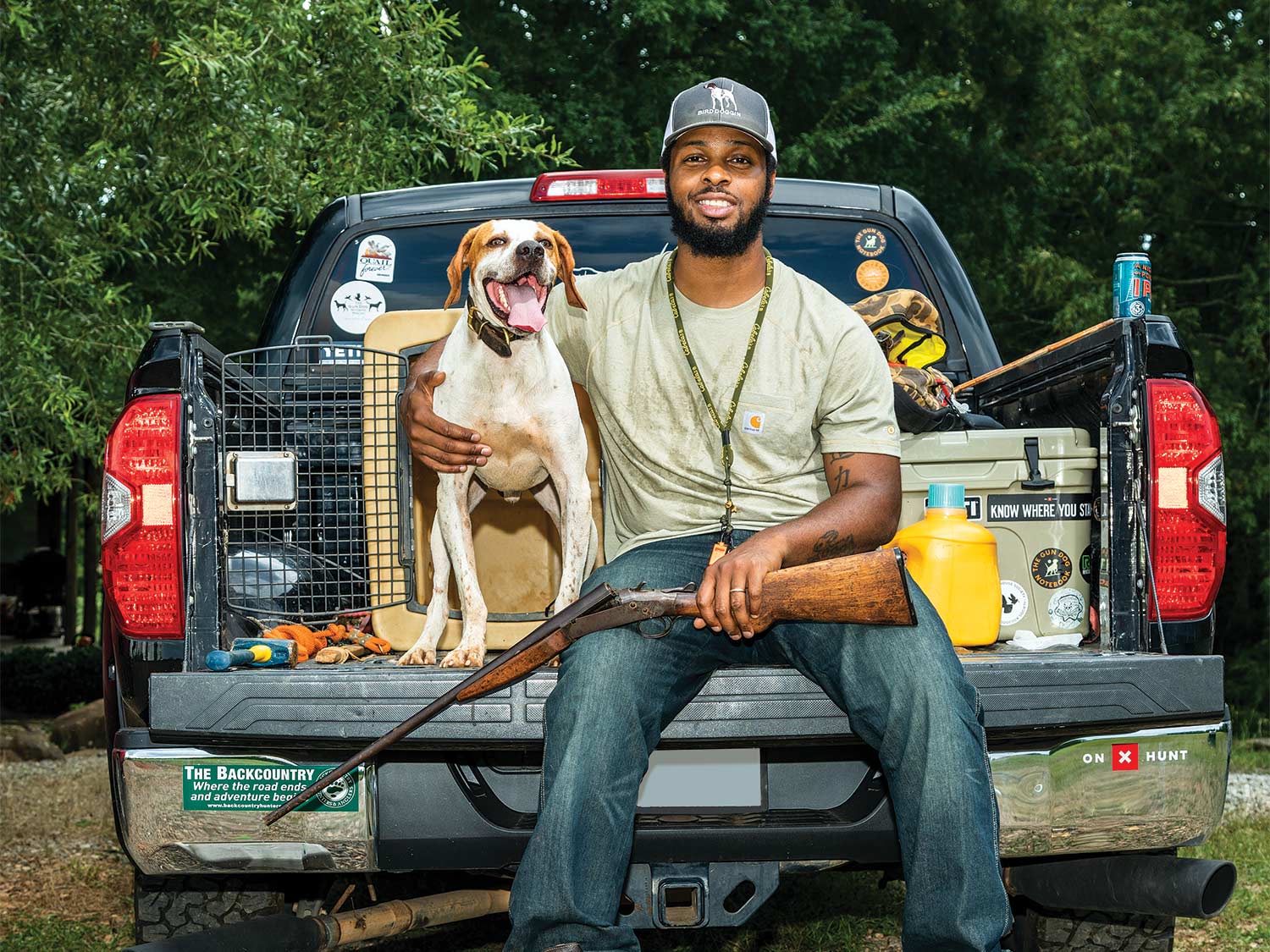 A hunter smiles, seated on a truck tailgate, with his hand wrapped around his dog.