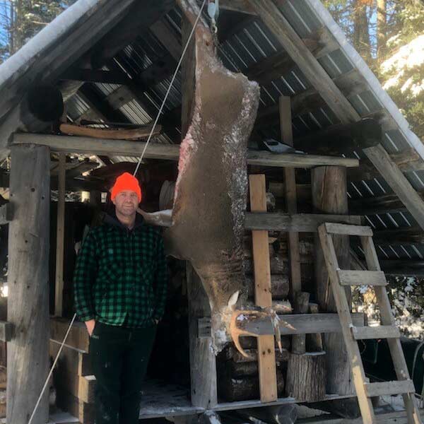 A hunter stands next to a buck hanging from a hunting camp hook.