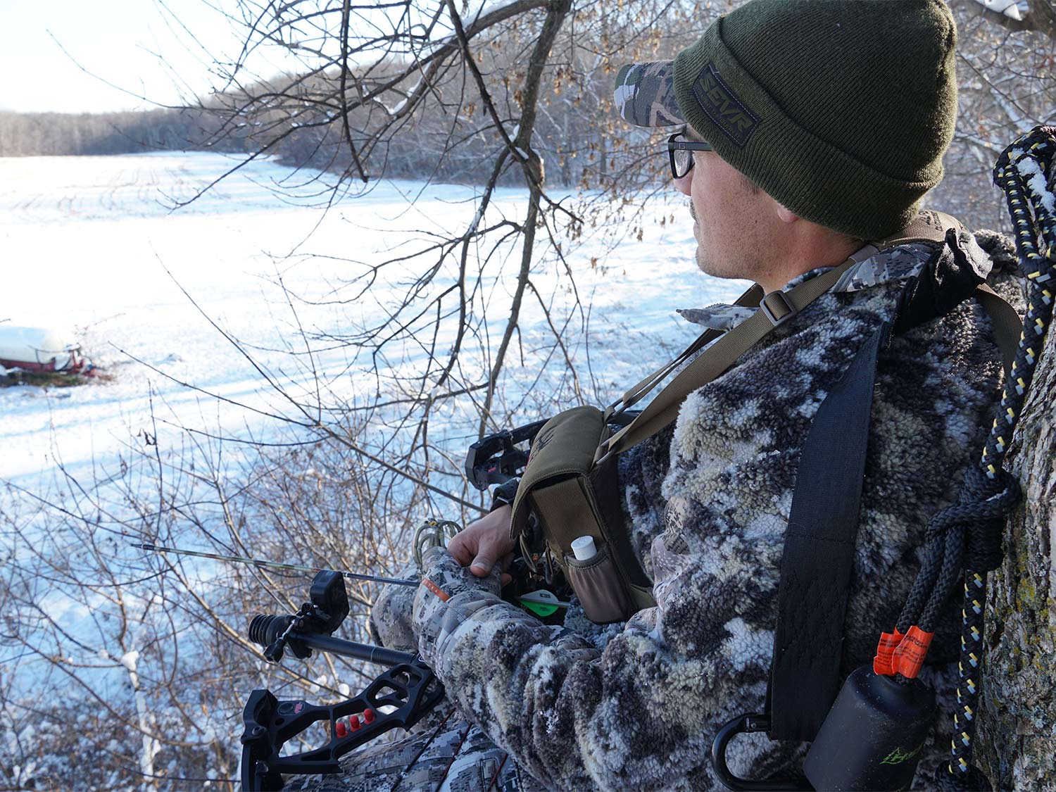 A hunter in full camo looks out over a field covered in snow.