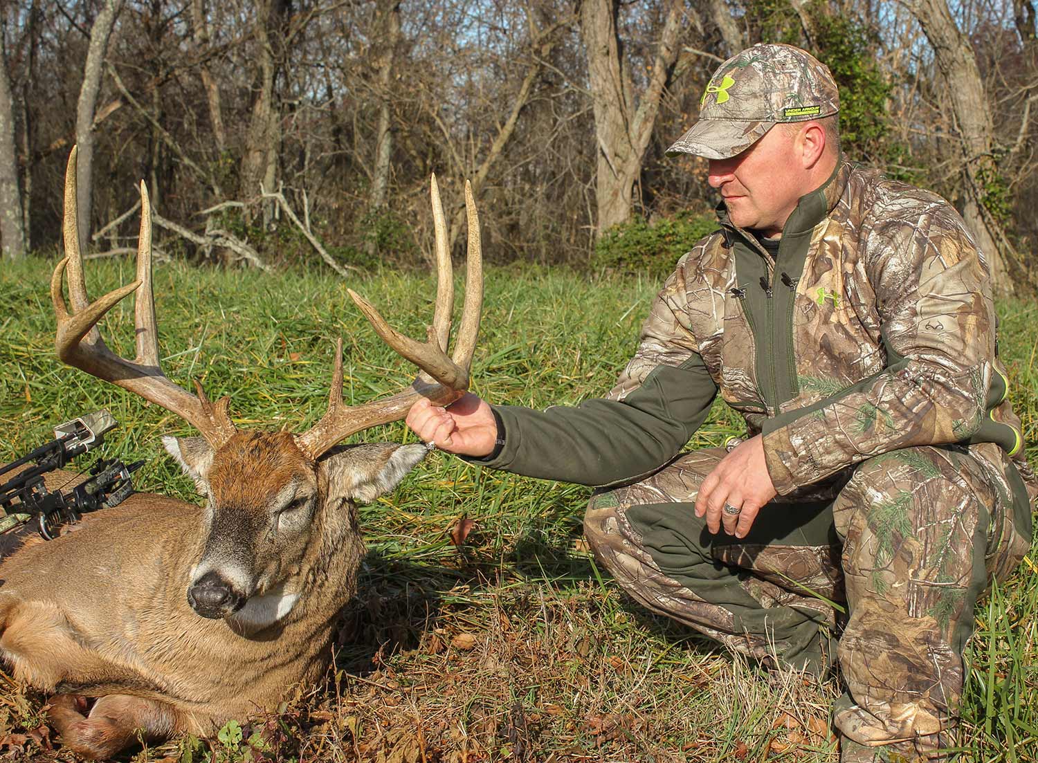 A hunter in full camo kneels beside a dropped whitetail buck and holds its head up by the antler.