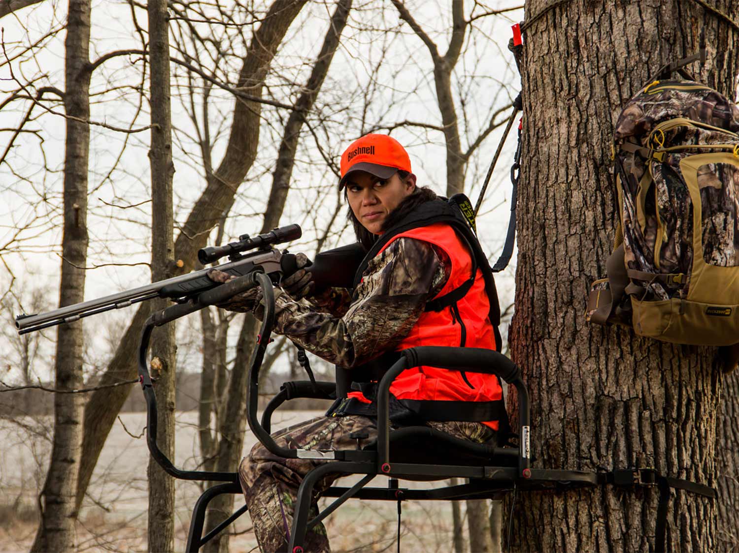 A woman hunter in full camo and an orange vest holds a scoped rifle while seated in a tree stand.