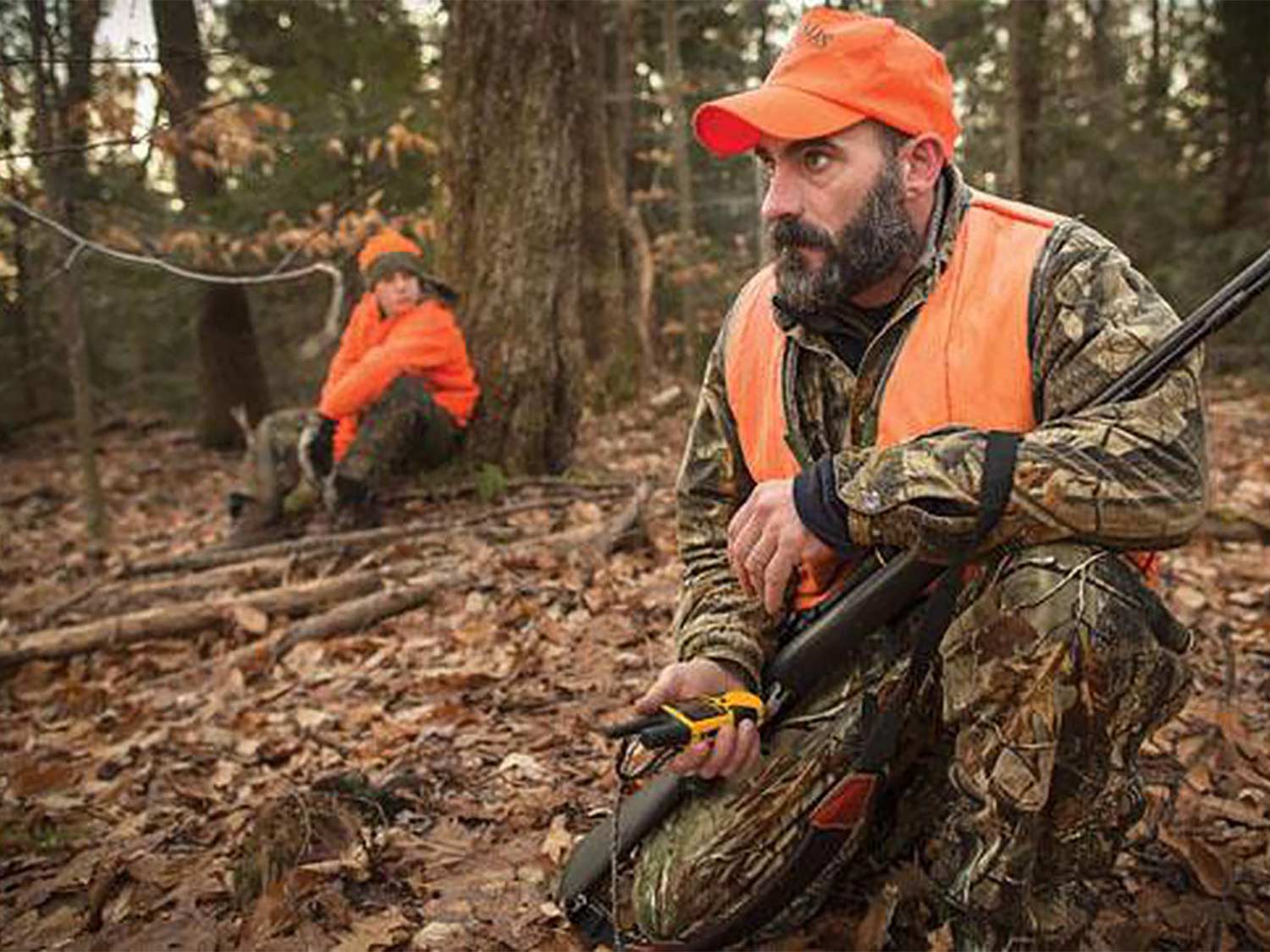 A hunter holds their rifle and crouches in the woods, clutching a 2-way radio in his hand.