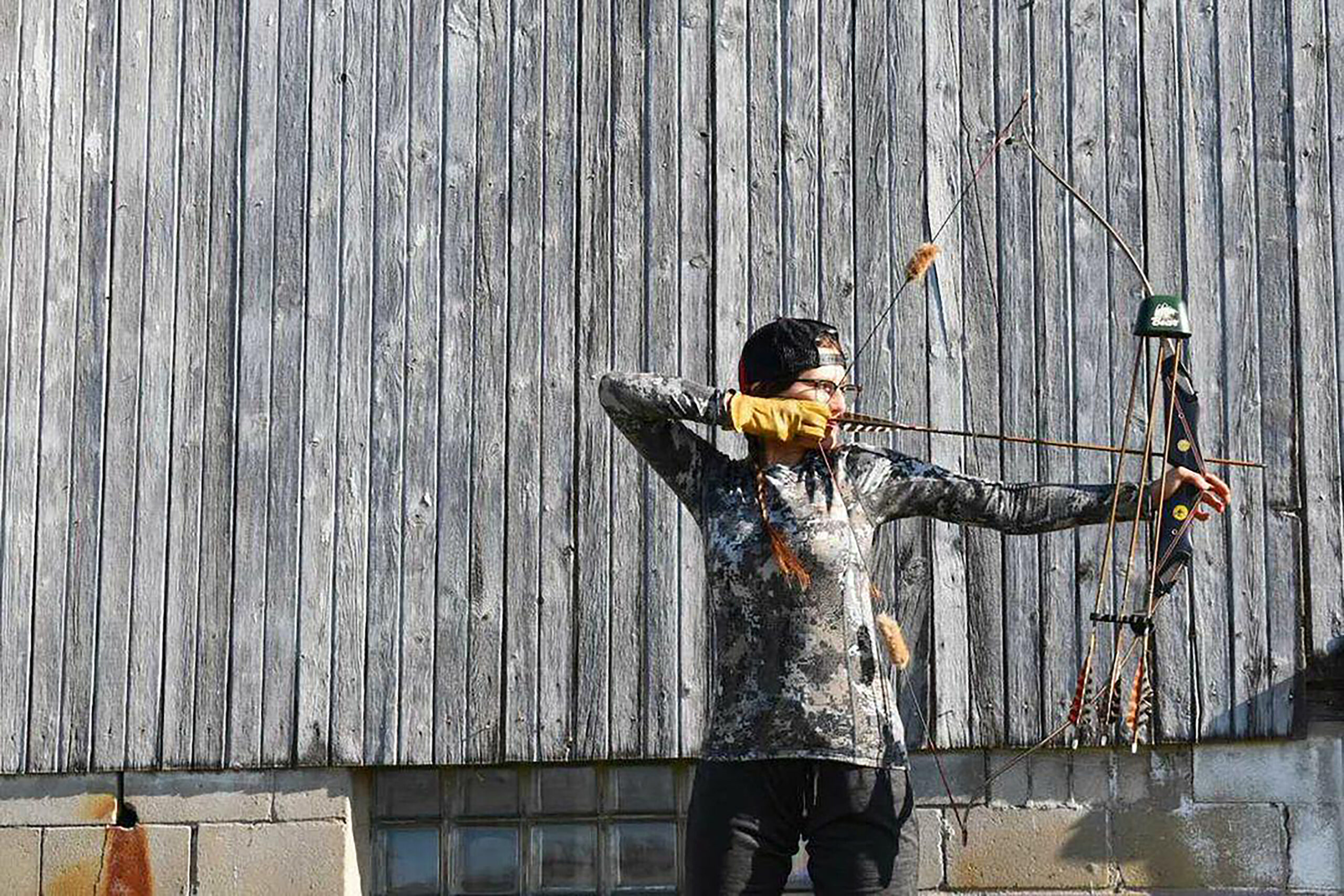 A female archer draws a traditional recurve bow in front of a wooden barn .