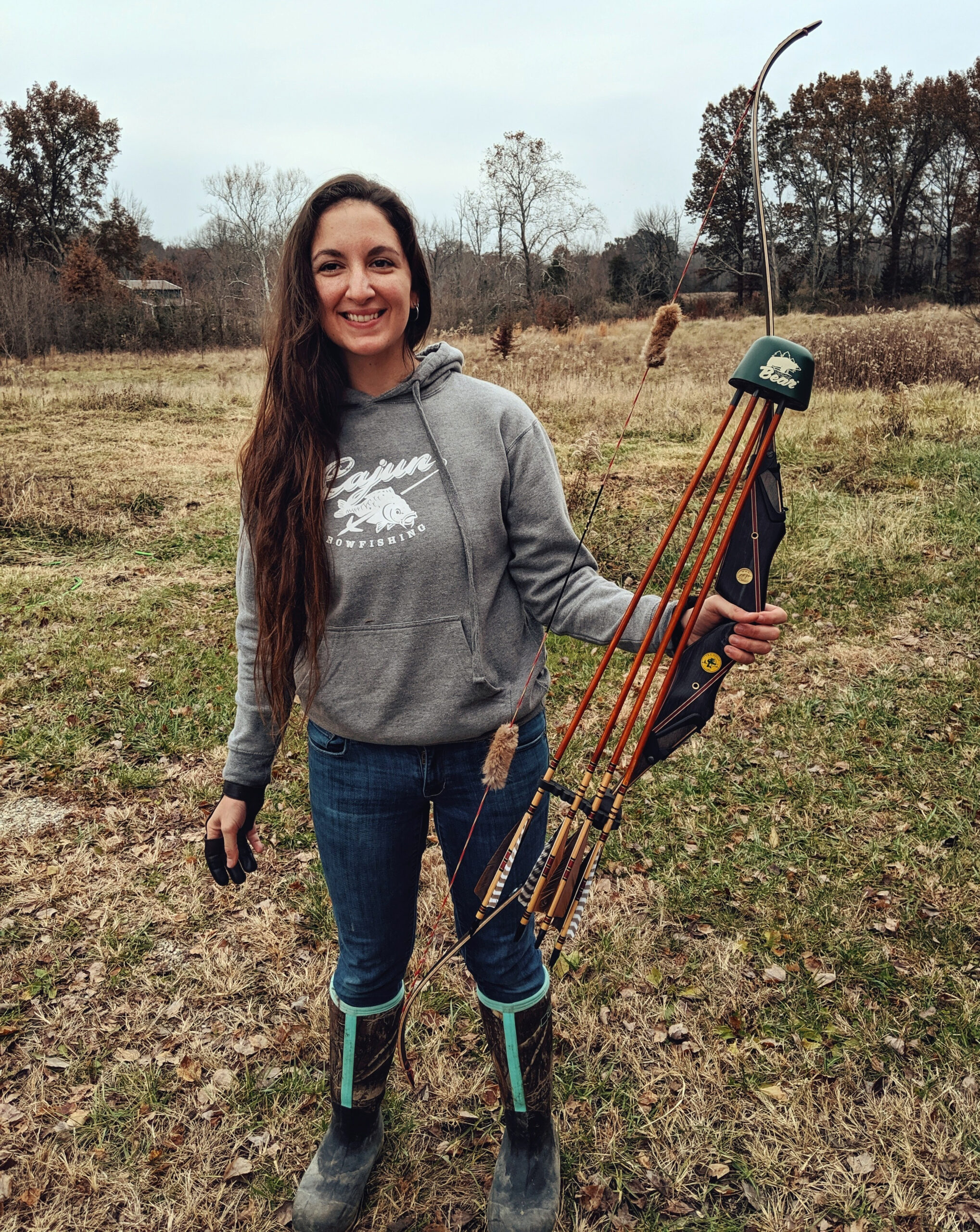 A woman with long hair in a gray hoodie holds a traditional recurve bow with a quiver full of arrows on a gray November day.