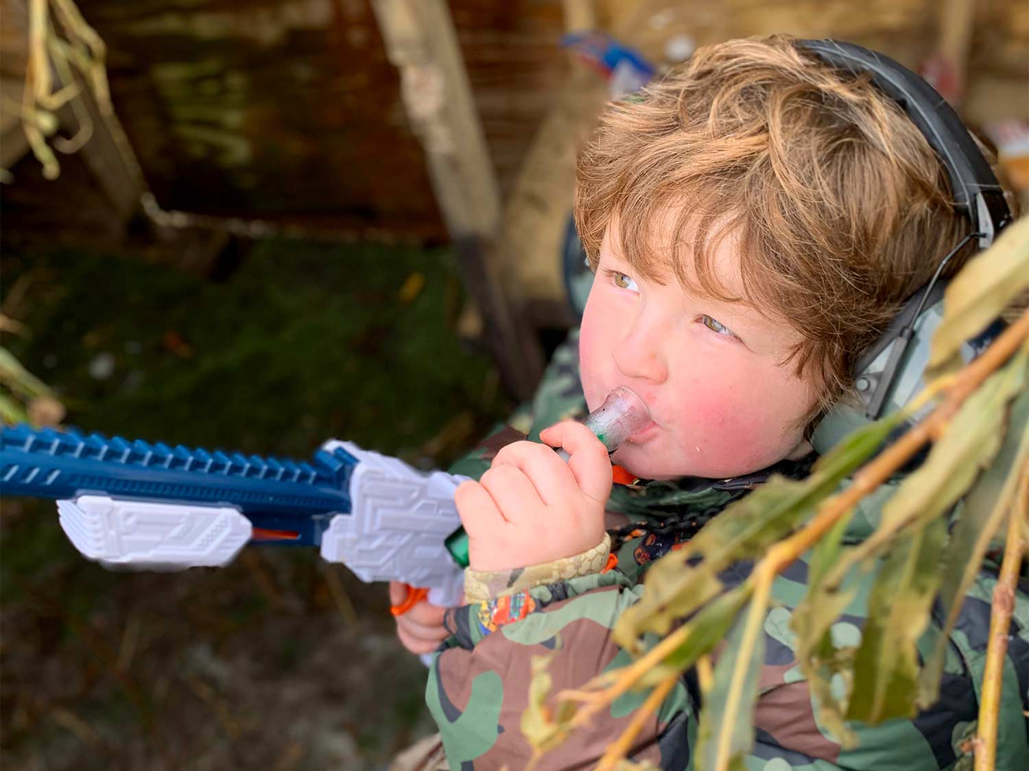 A young kid blows on a duck call while holding a toy gun.