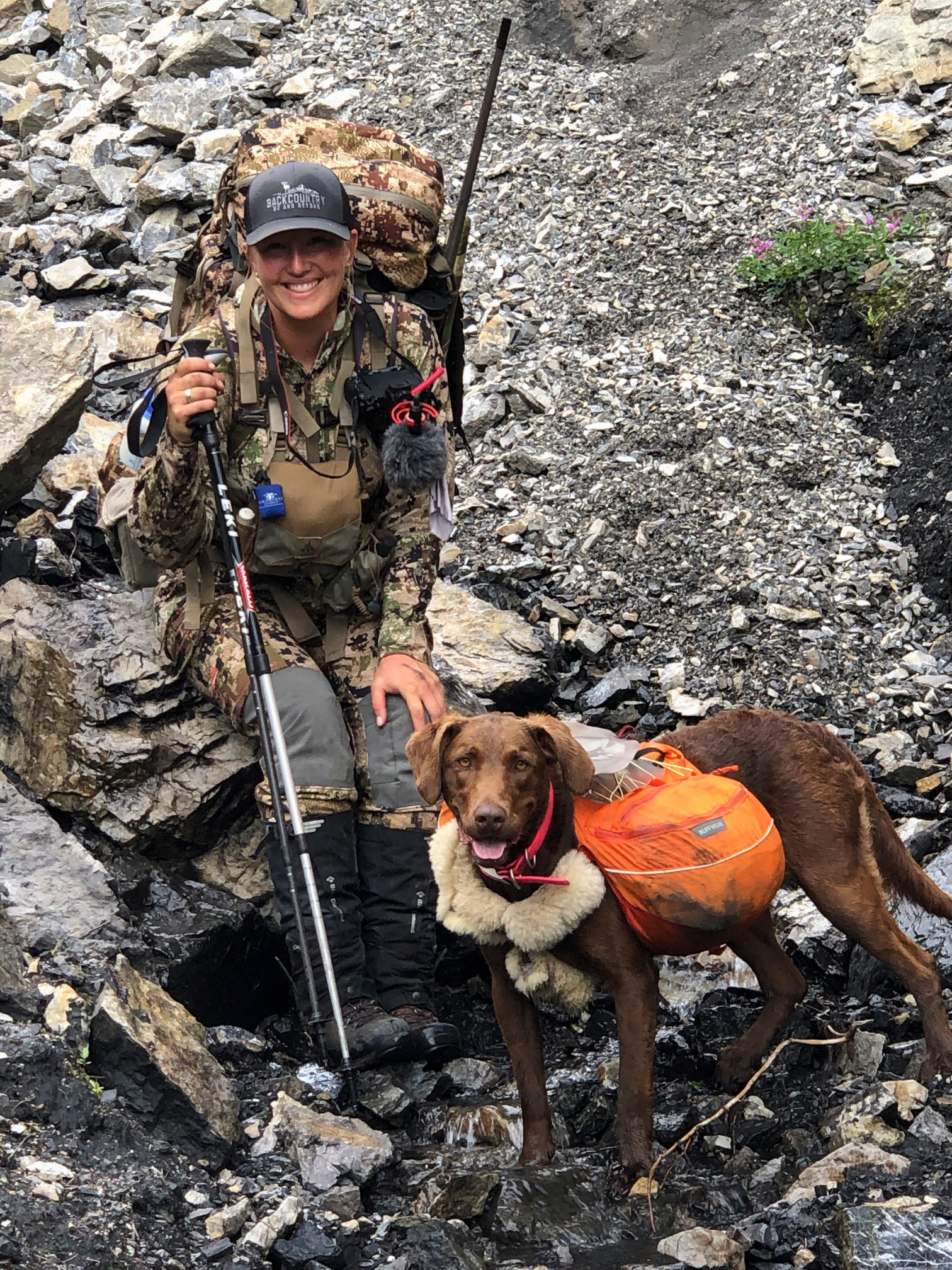 A female big-game guide with trekking poles and a heavy pack rests on a rock beside her dog.