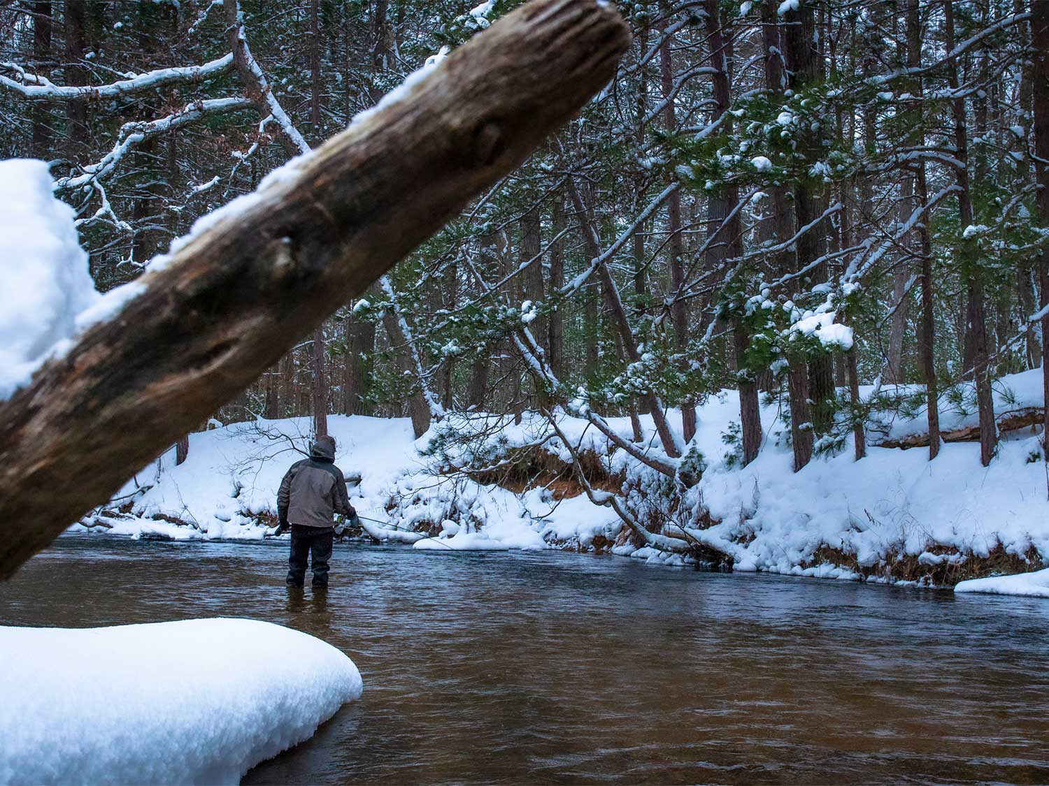 An angler wades into a river and fishes in the snow.