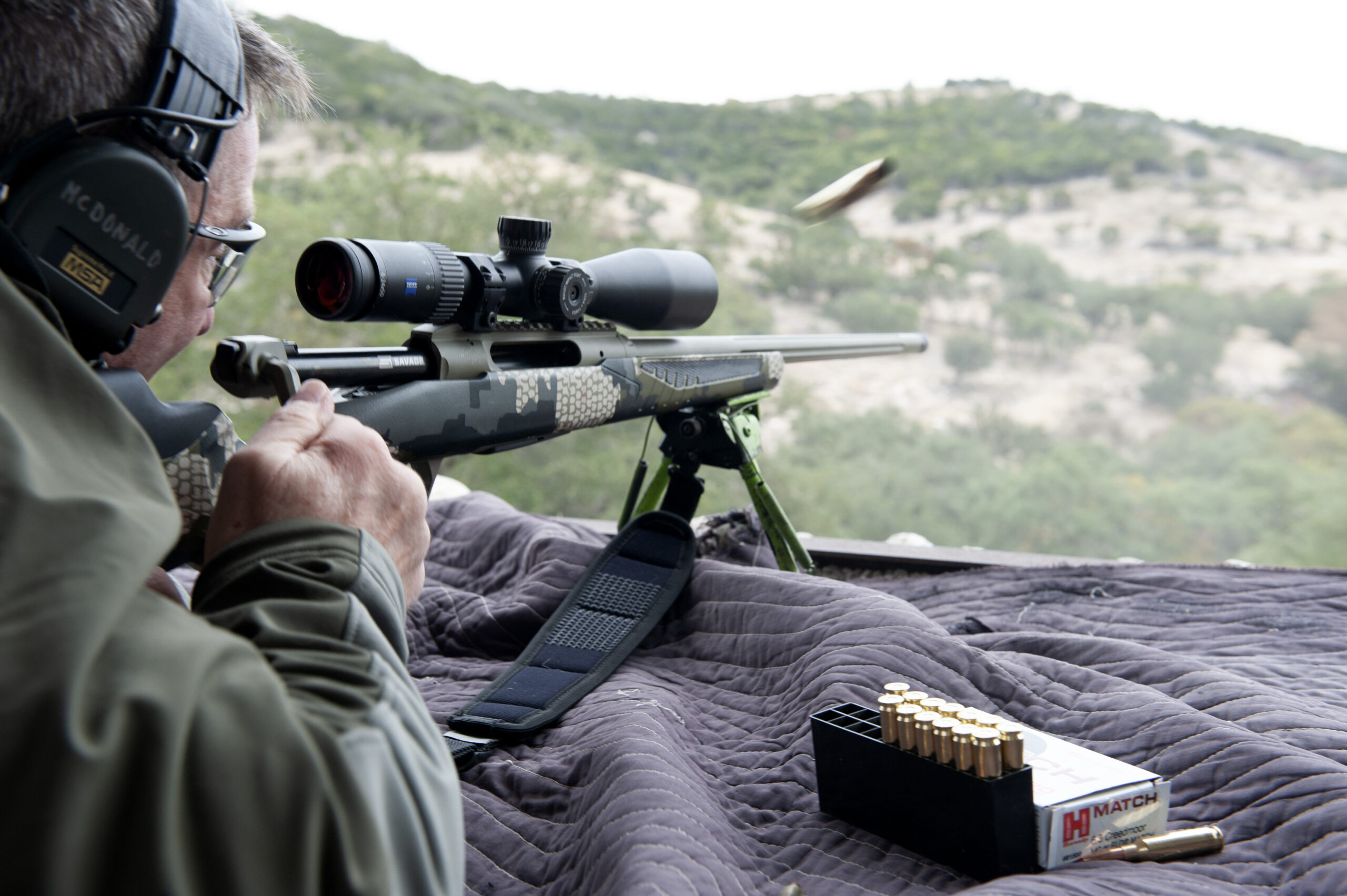 A man cycles a straight-pull rifle action, ejecting a brass cartridge while shooting prone.