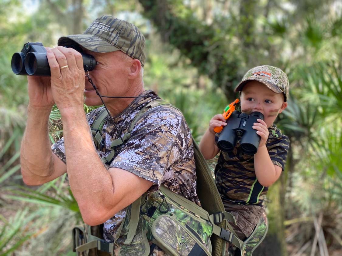 A hunter and child in the woods using binoculars.